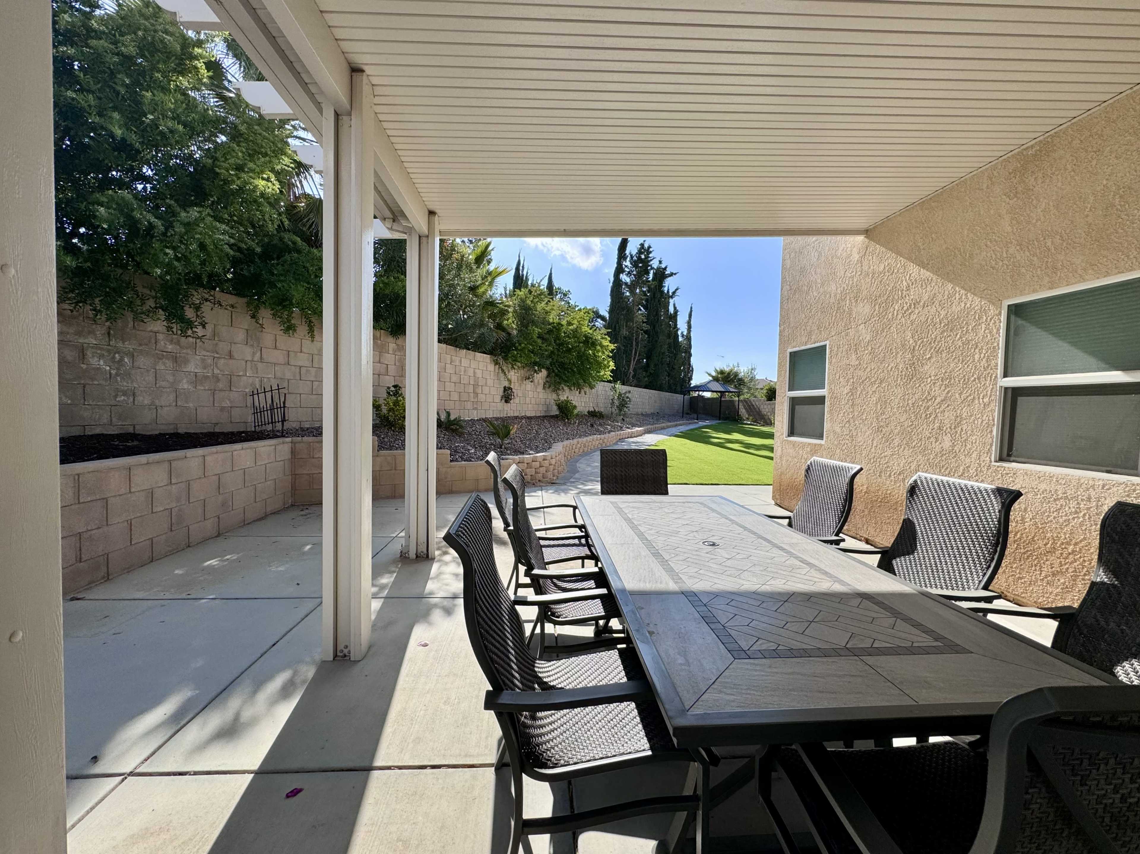 A shaded outdoor dining area features a large table surrounded by black chairs, overlooking a grassy yard with shrubs and trees in the background.