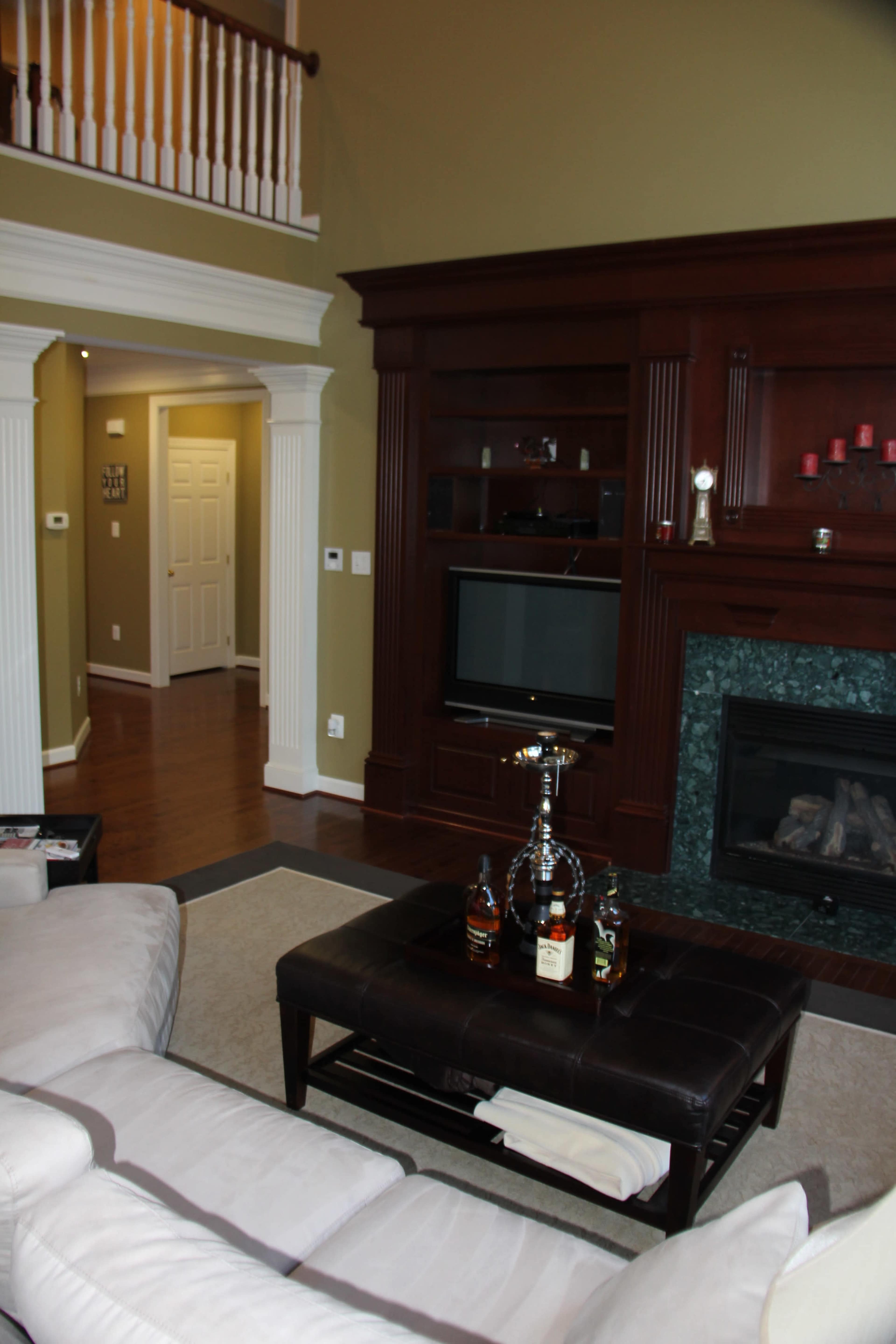 A living room with a dark wooden entertainment center, a fireplace, and a coffee table featuring an arrangement of bottles, with a staircase and hallway visible in the background.