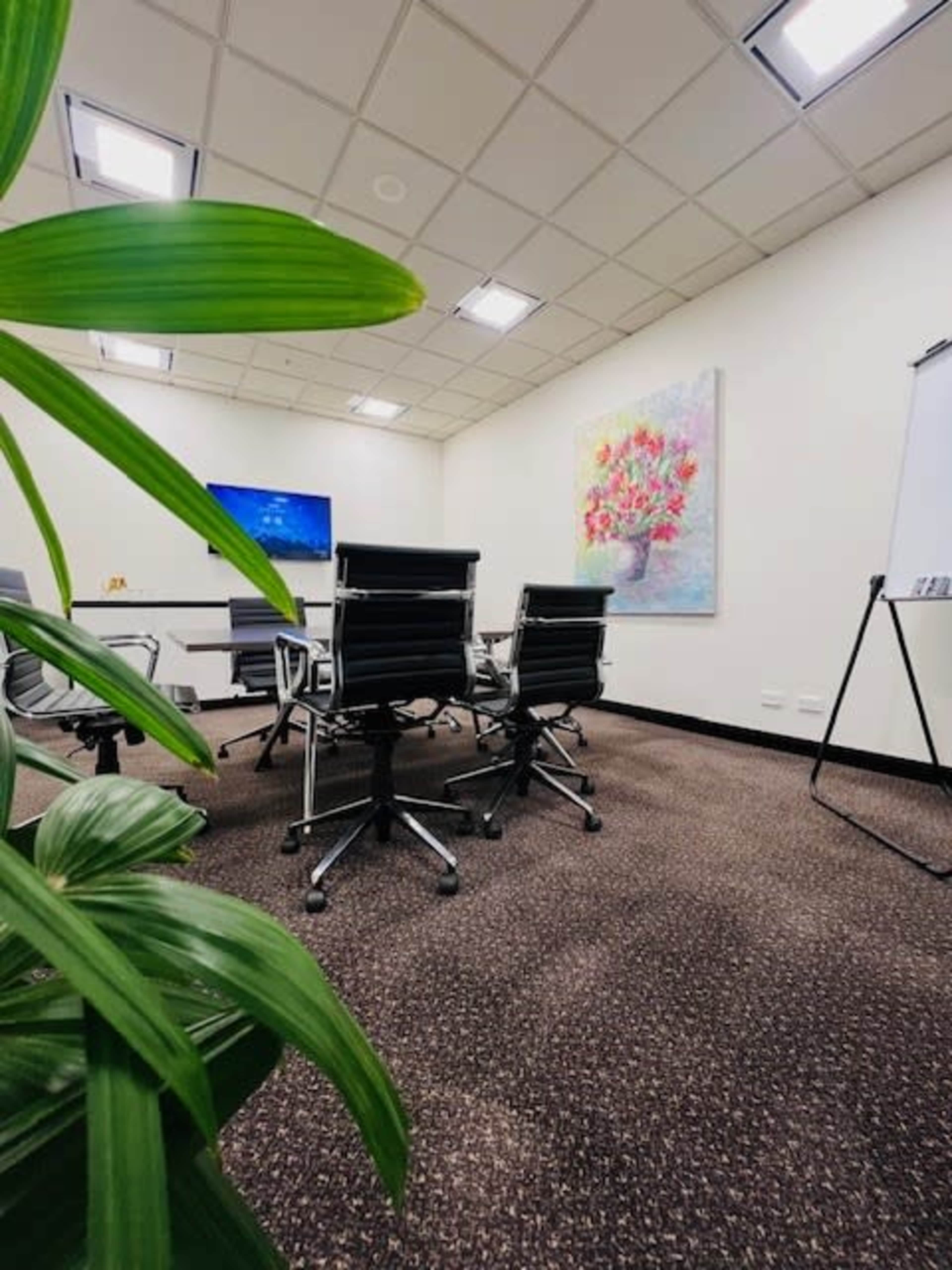 A modern conference room features sleek black chairs around a round table, with a large floral painting on the wall and a screen mounted in the corner.