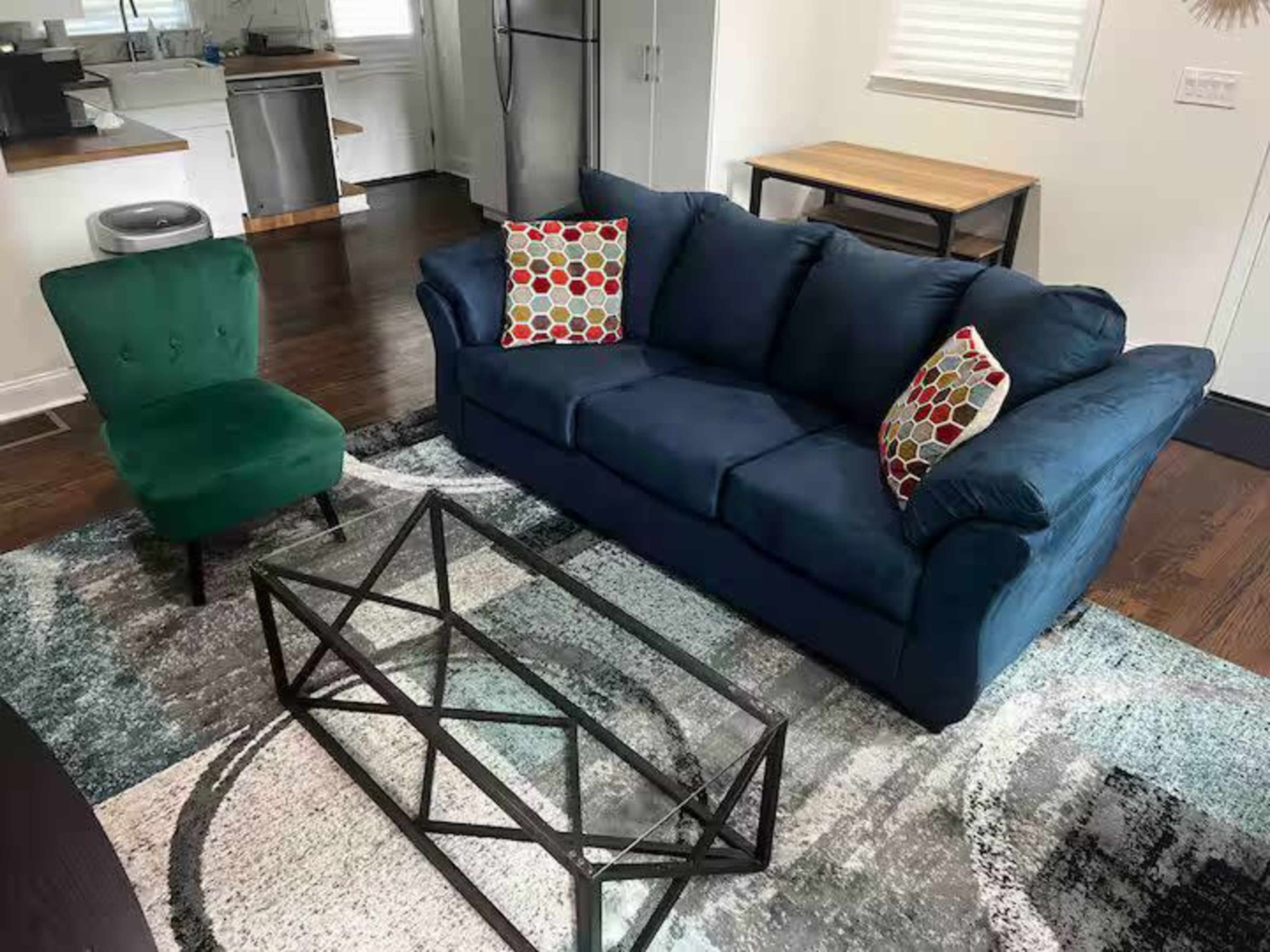 The image shows a living room featuring a blue couch with decorative pillows, a green chair, a geometric coffee table, and a patterned area rug.