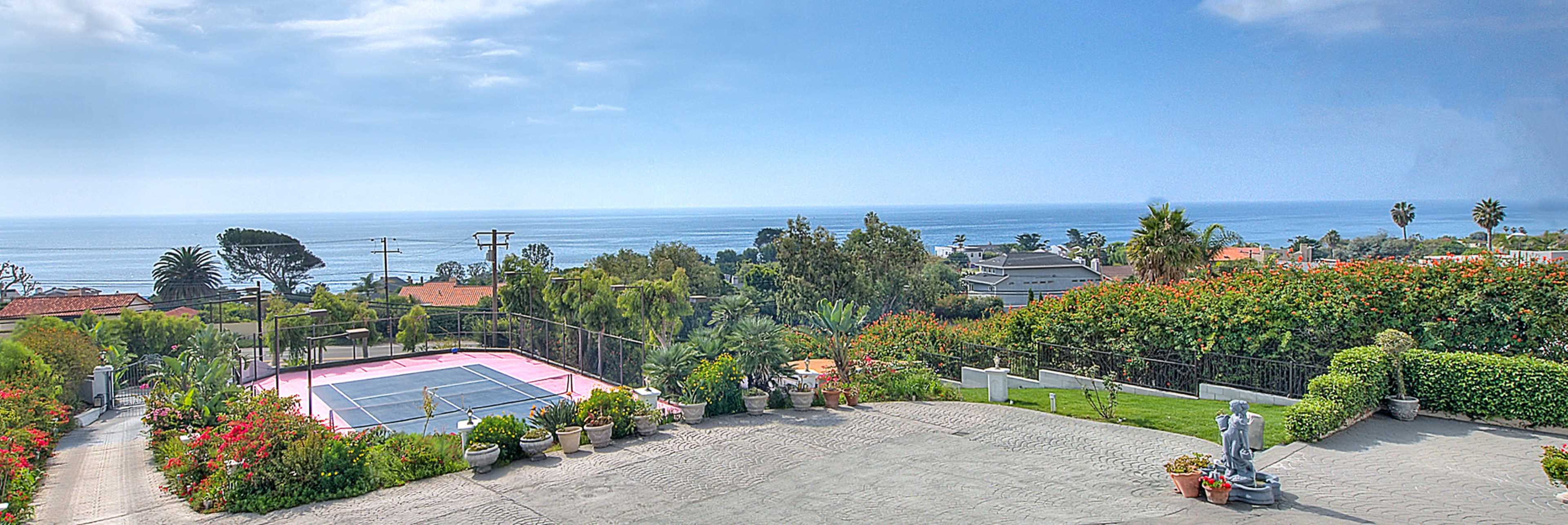 A coastal view featuring a pink tennis court surrounded by lush greenery and blooming flowers, with the ocean visible in the background.