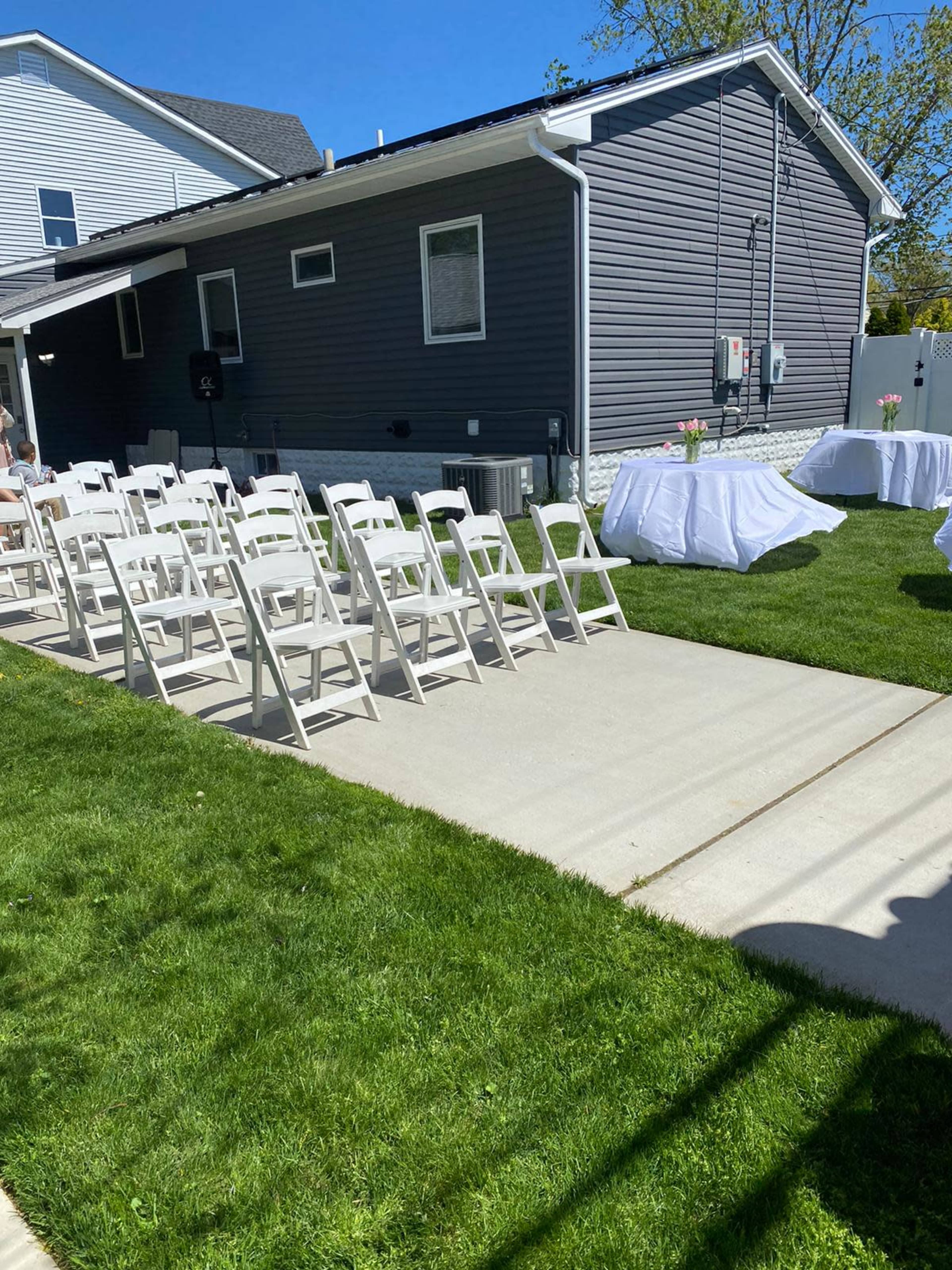 A row of white folding chairs is neatly arranged on a concrete path beside a grassy area next to a gray house, with a table covered in white cloth in the background.