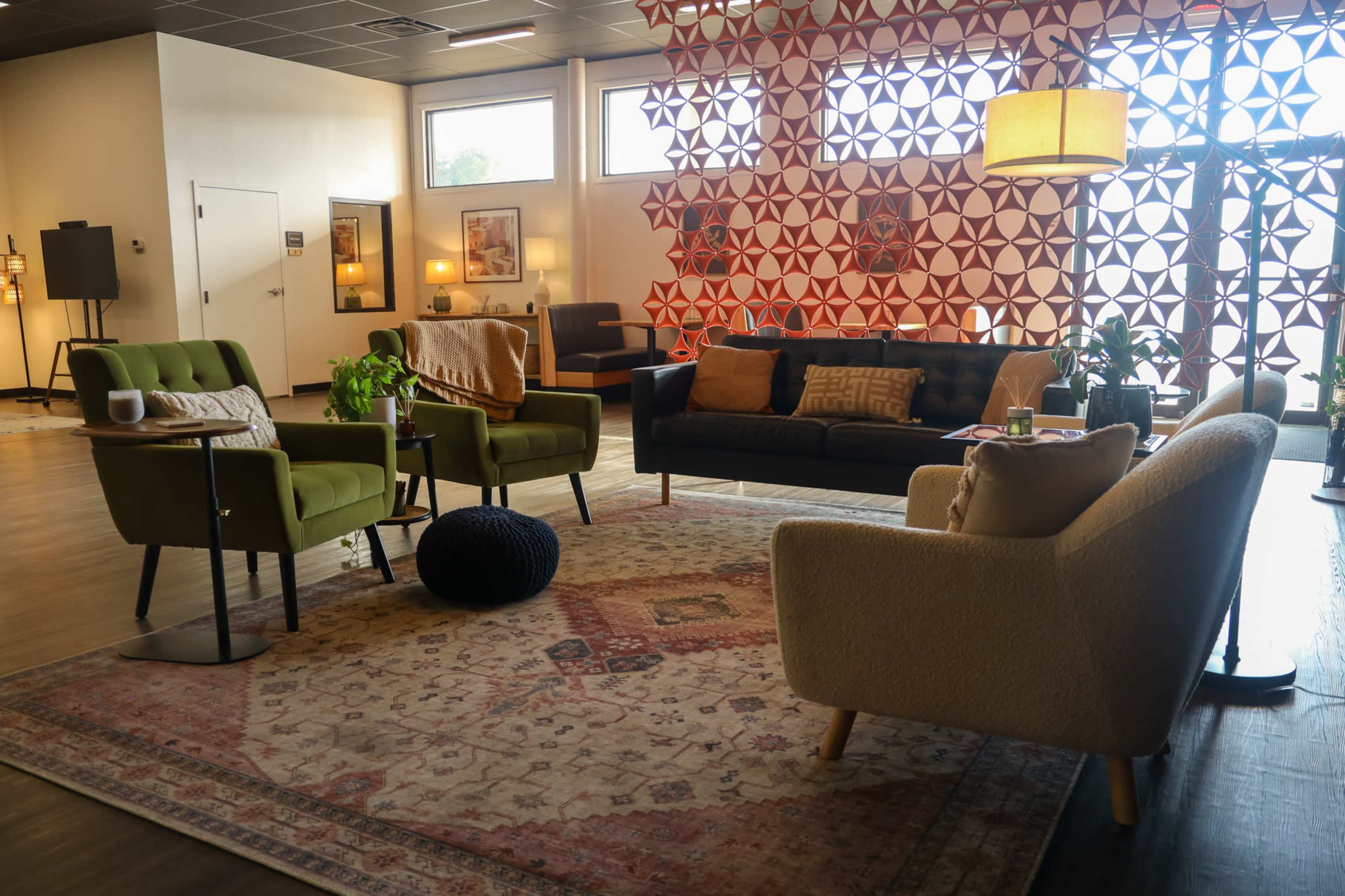 The image depicts a modern lounge area featuring various seating options, a patterned backdrop, and decorative plants on a large rug.