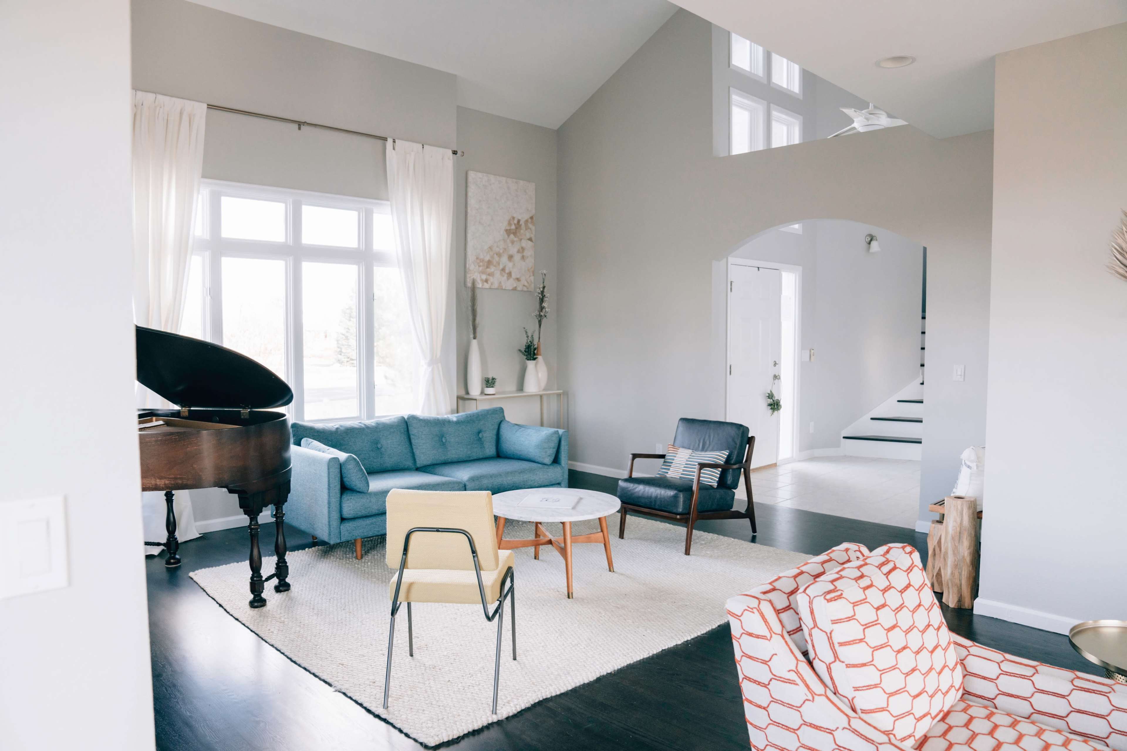 The image shows a modern living room with a blue couch, a grand piano, a round coffee table, and various seating options arranged around a light-colored area rug.