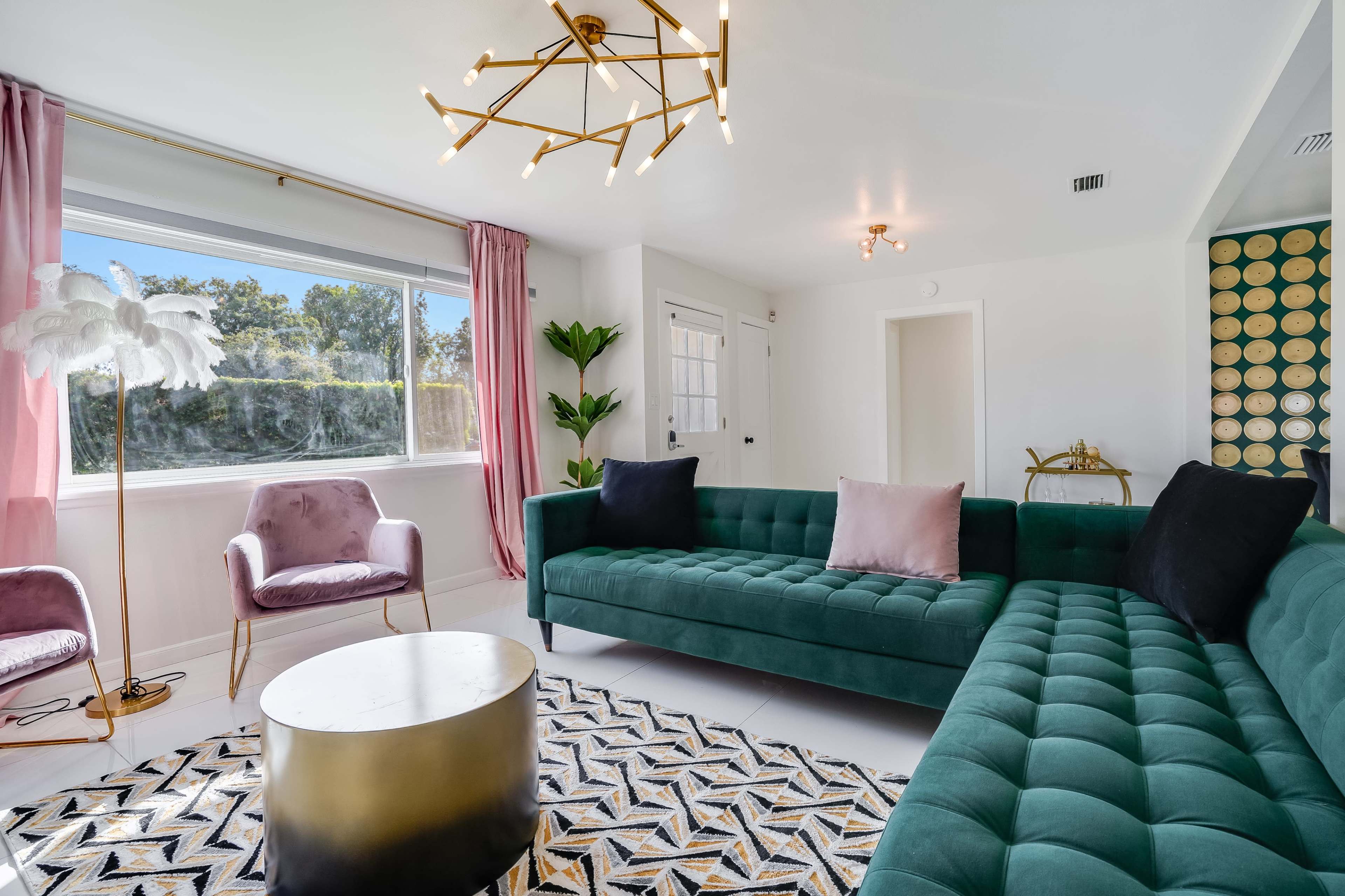 A modern living room featuring a green tufted sofa, a round gold coffee table, a pink velvet armchair, and large windows with sheer curtains.