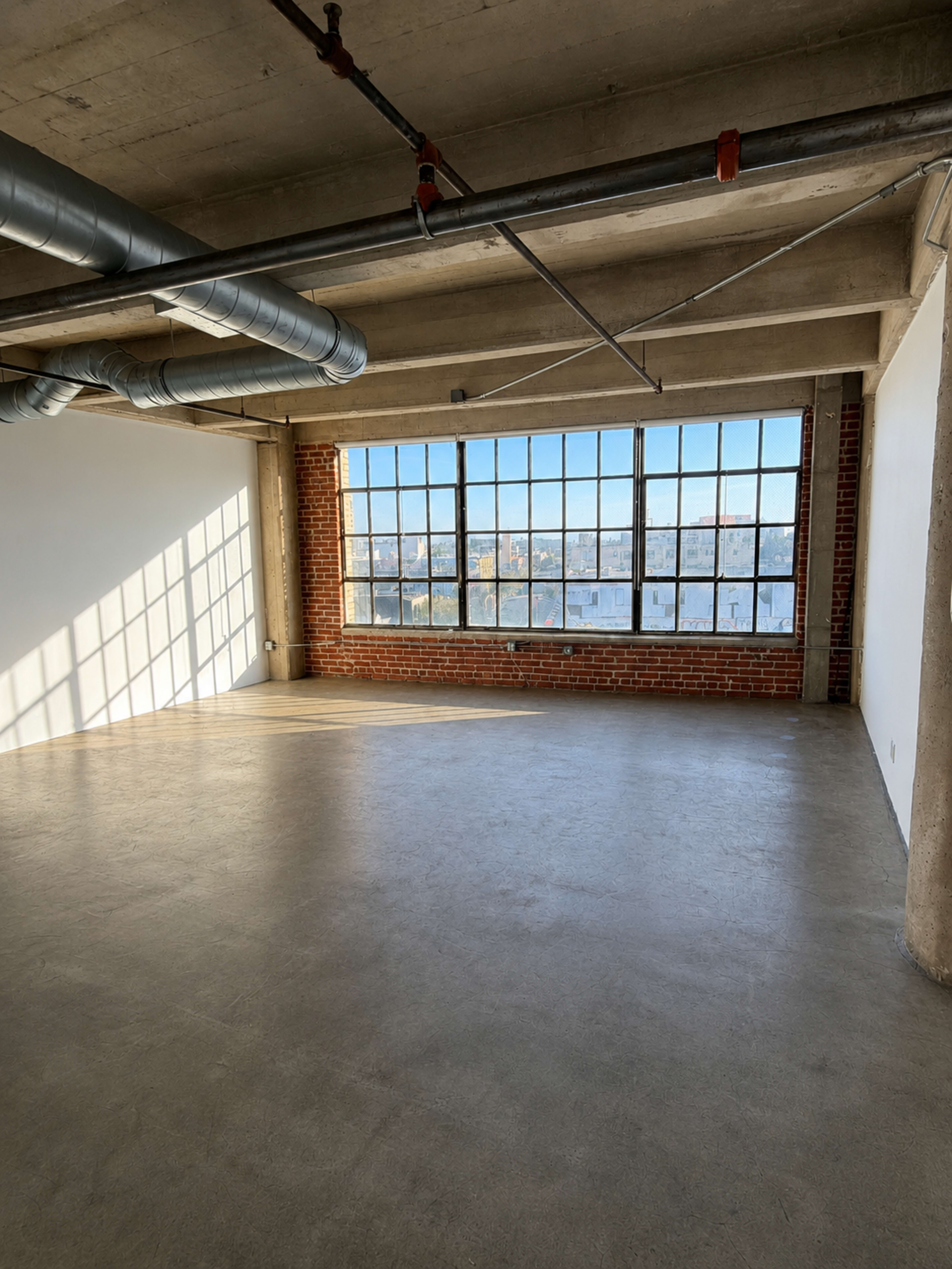 The image shows an industrial-style room with large windows allowing natural light to enter, exposing brick walls and concrete flooring.