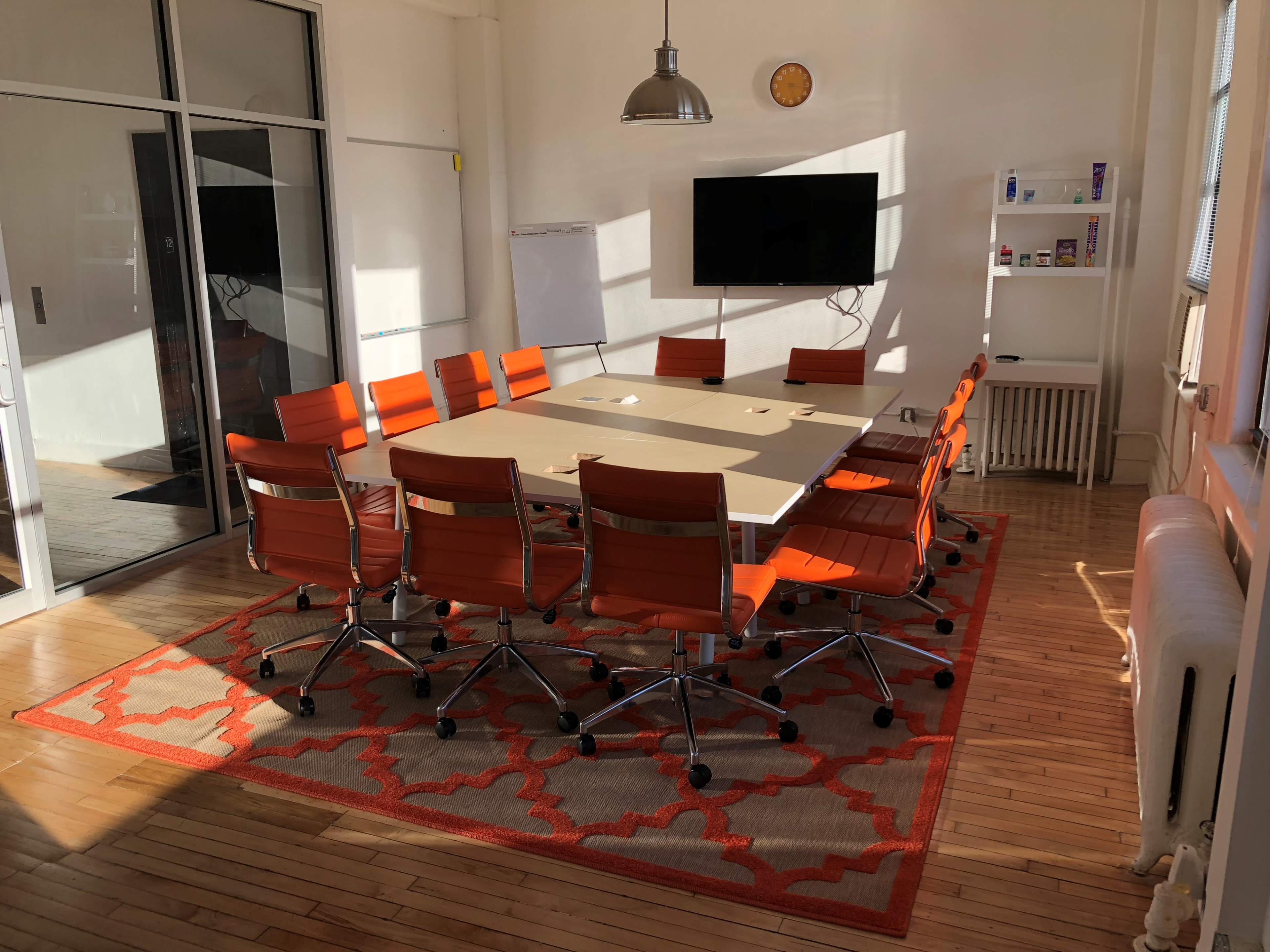 A conference room features a large table surrounded by orange chairs, with a wall-mounted screen and a whiteboard visible.