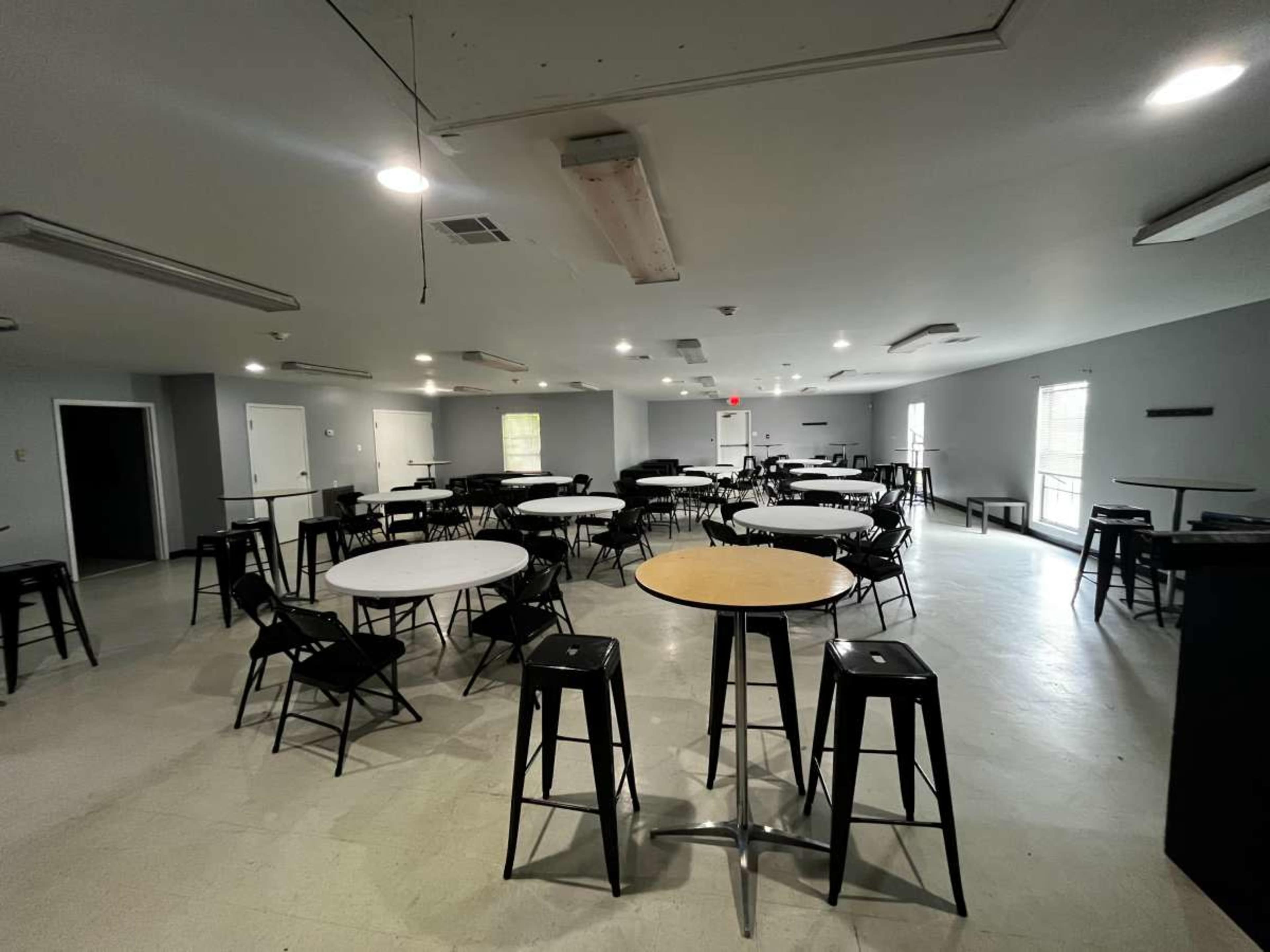 The image shows a spacious room with several round and square tables, surrounded by black stools, and illuminated by overhead lights.