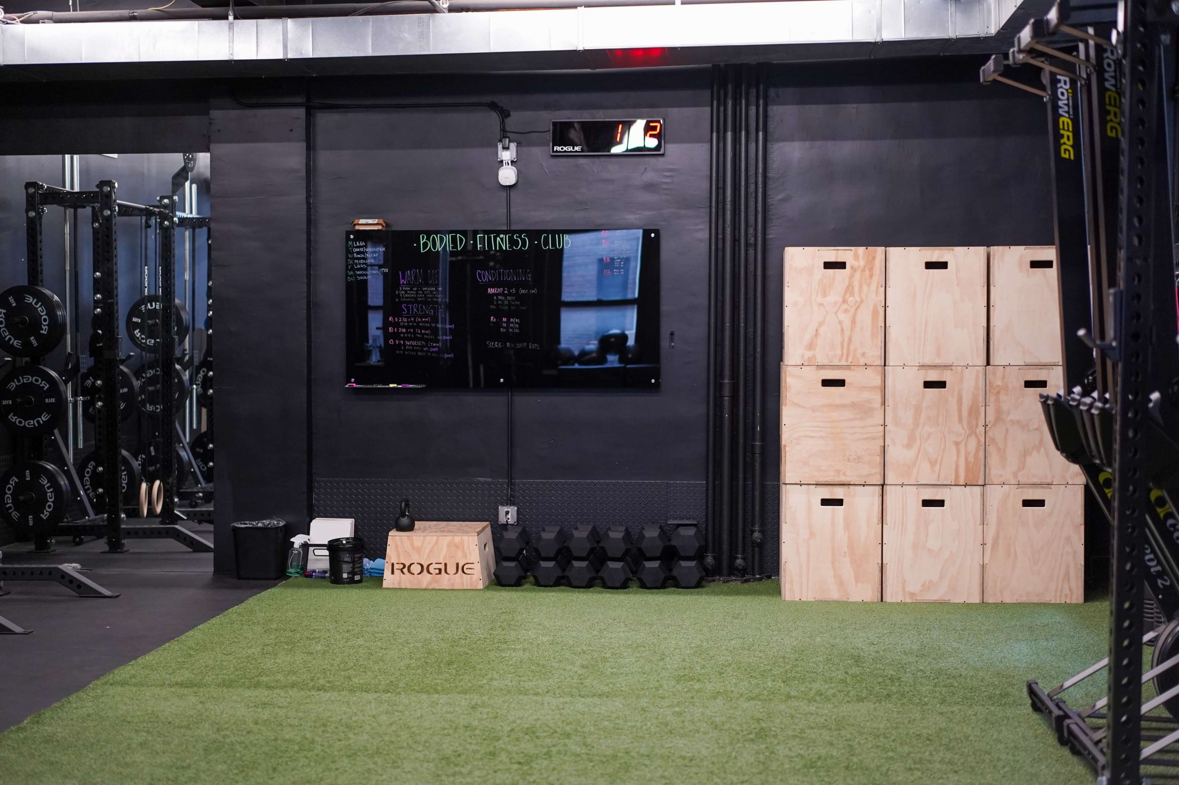 A gym interior featuring a television mounted on a black wall, wooden storage boxes, dumbbells, and a green turf area.
