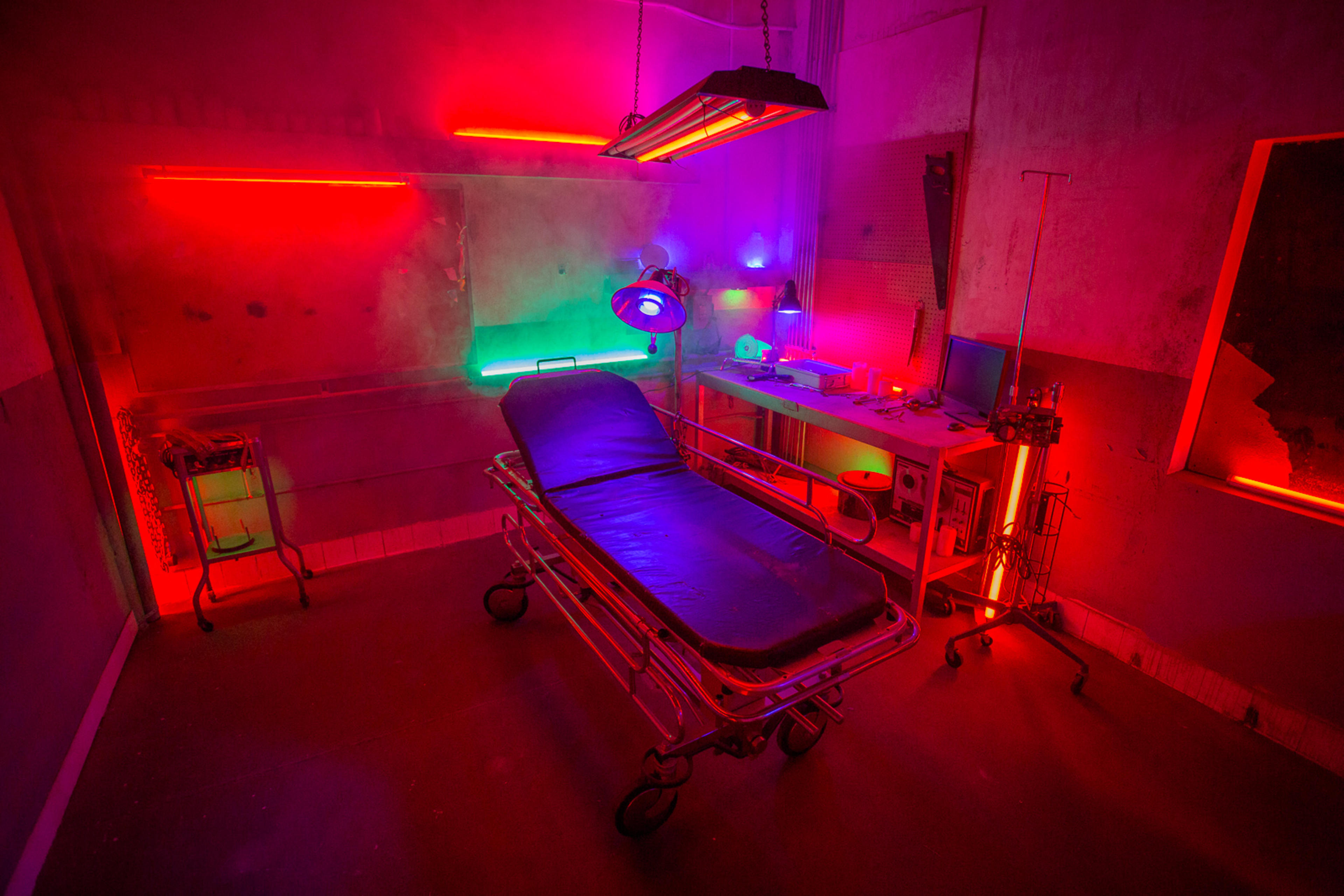 A dimly lit hospital room with neon lights illuminating a metal hospital bed, a medical lamp, and various equipment on a sleek table.