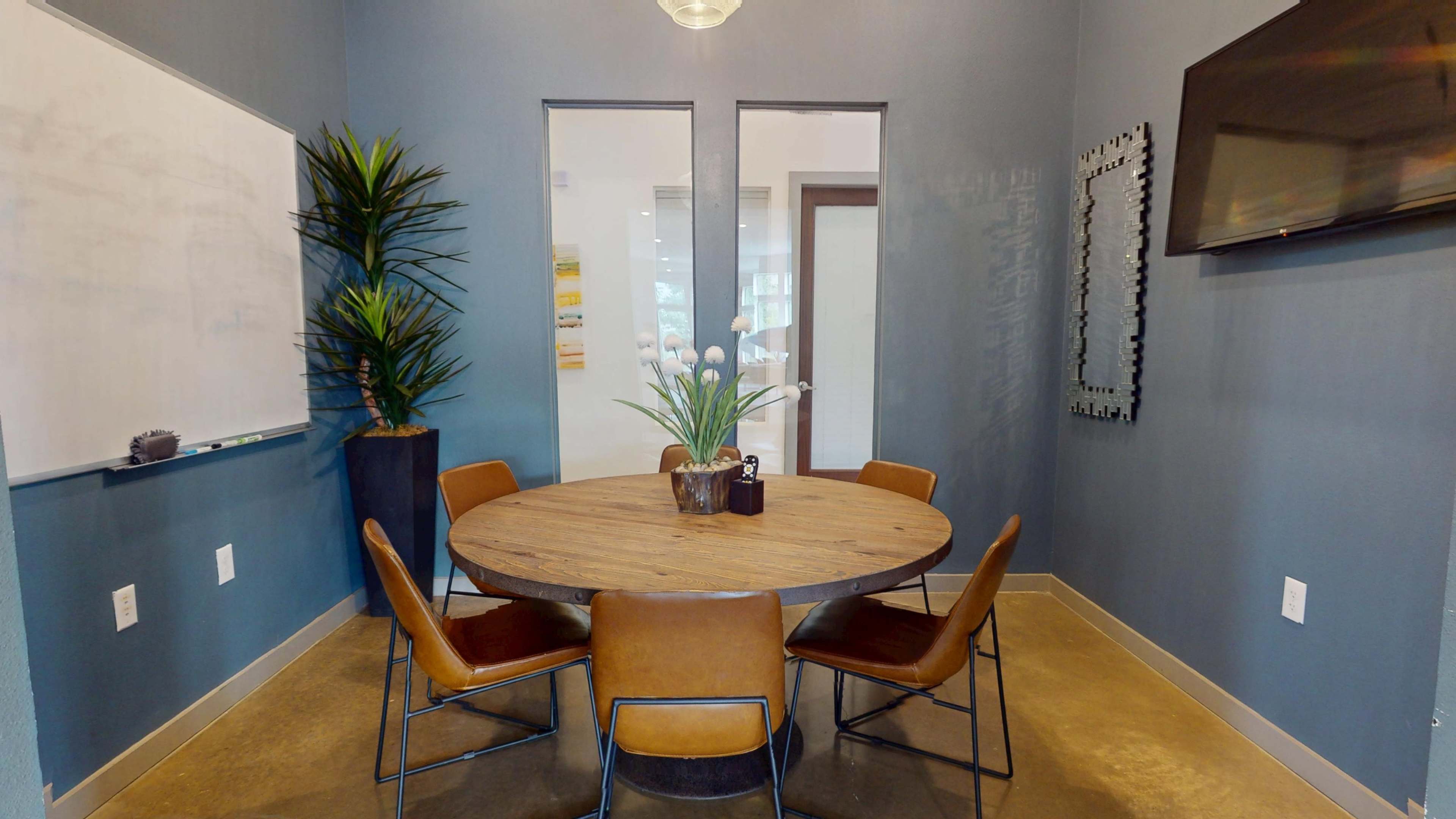 A conference room features a round wooden table surrounded by five chairs, with a plant and a whiteboard against the walls.