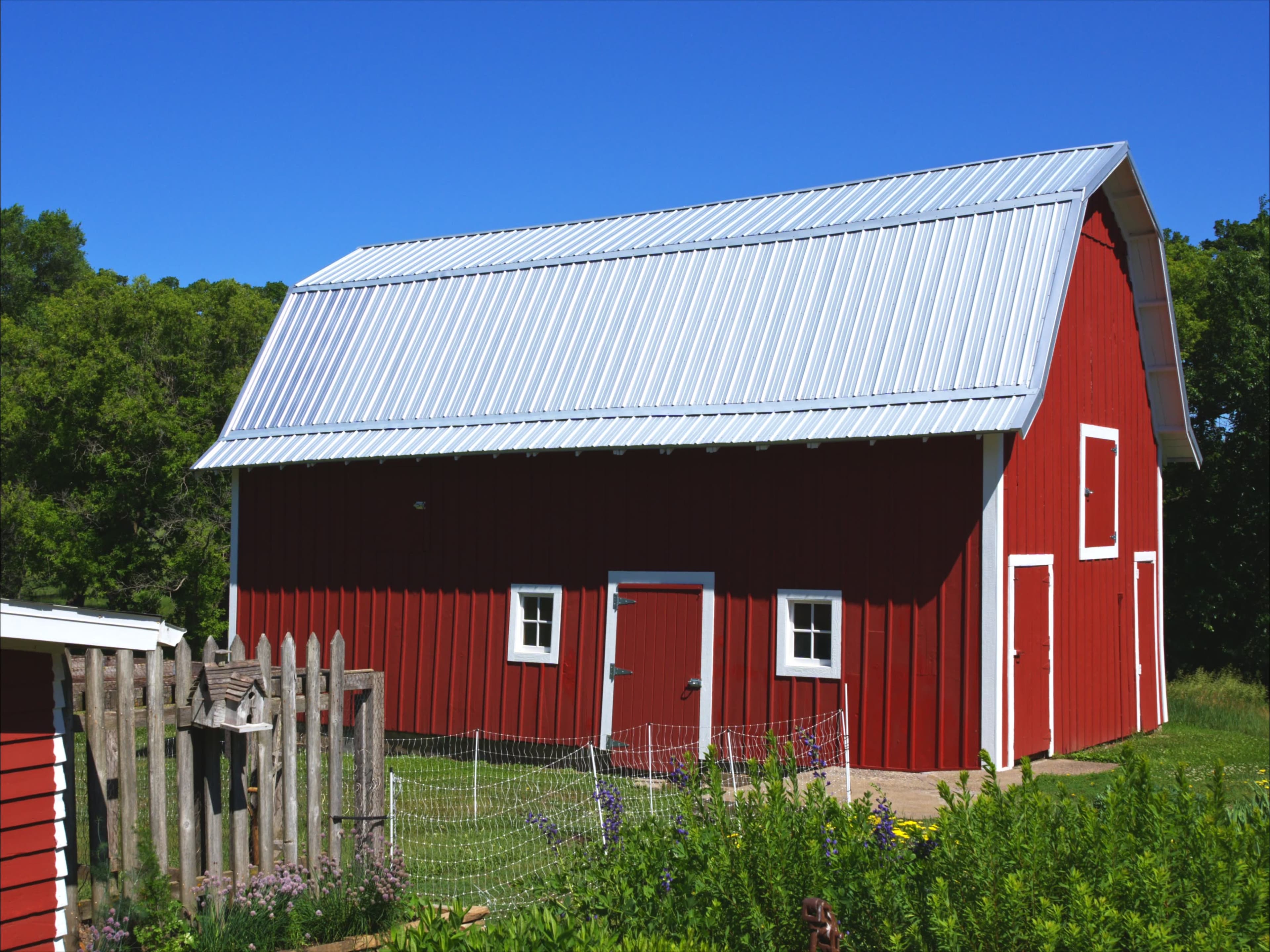 A red barn with a metal roof stands beside a wooden fence and lush greenery under a clear blue sky.