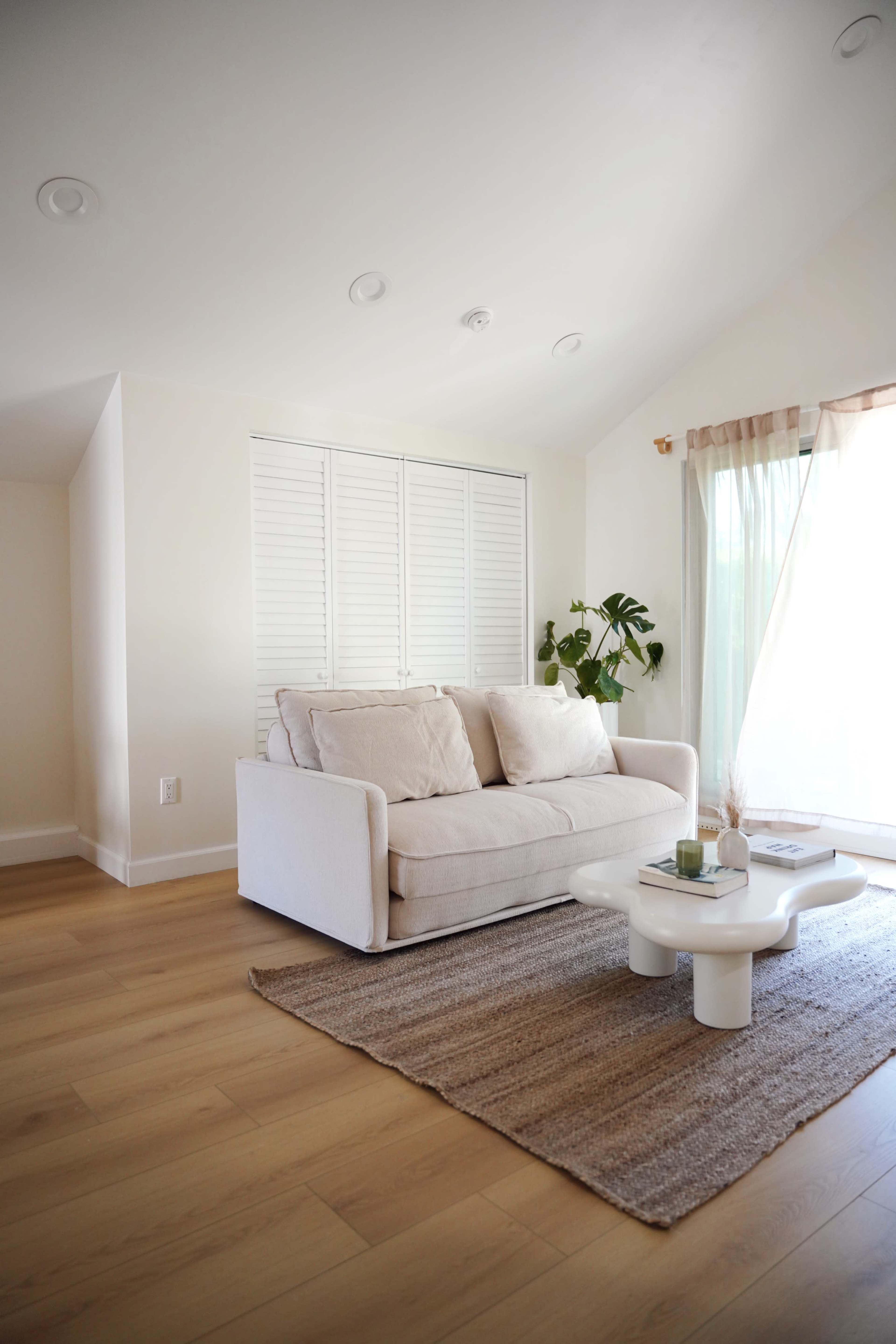 A light-filled living room features a beige sofa, a coffee table, and a large window with sheer curtains.