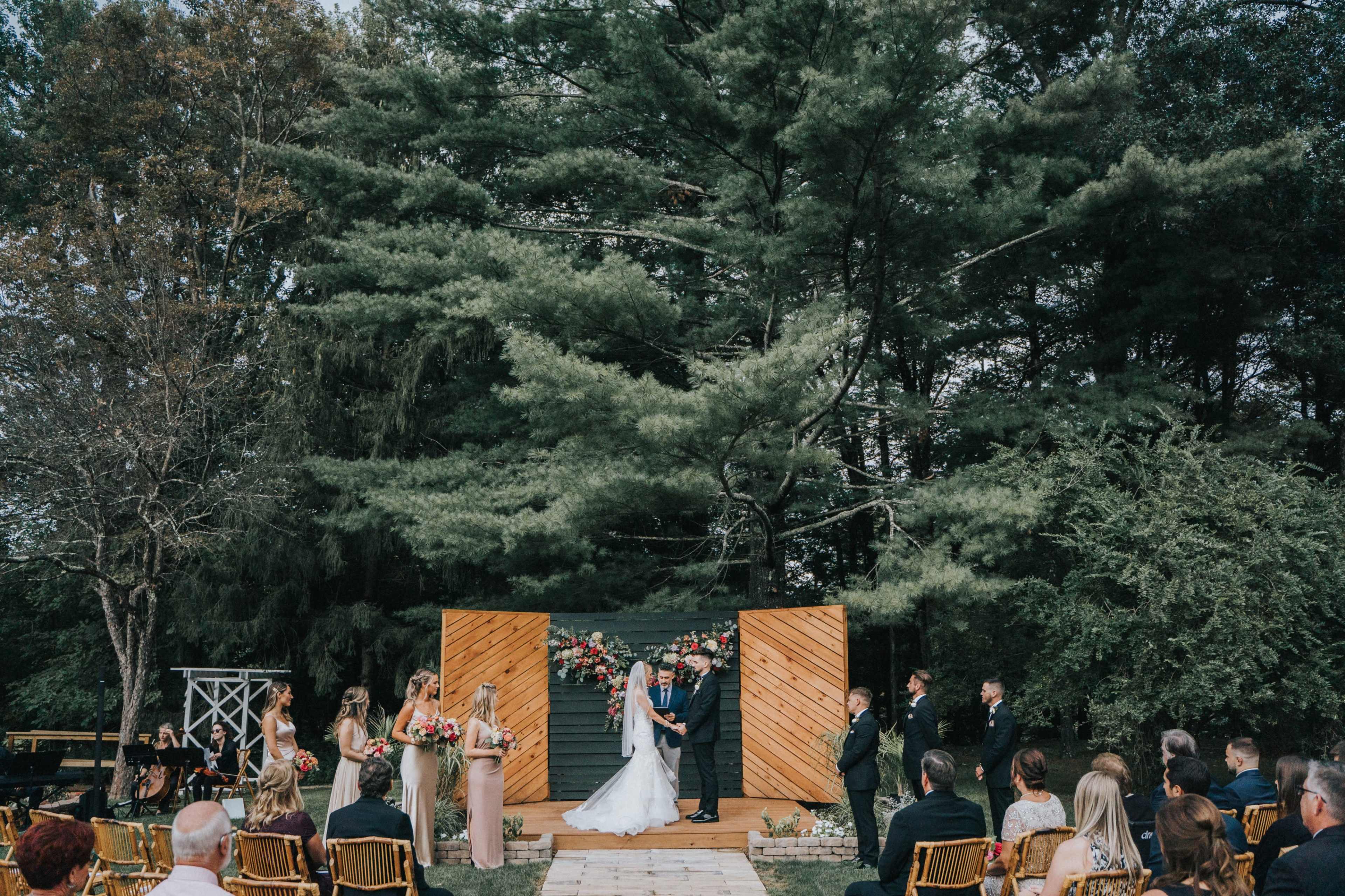 A wedding ceremony takes place outdoors on a wooden platform, surrounded by guests seated in rows and flanked by trees.