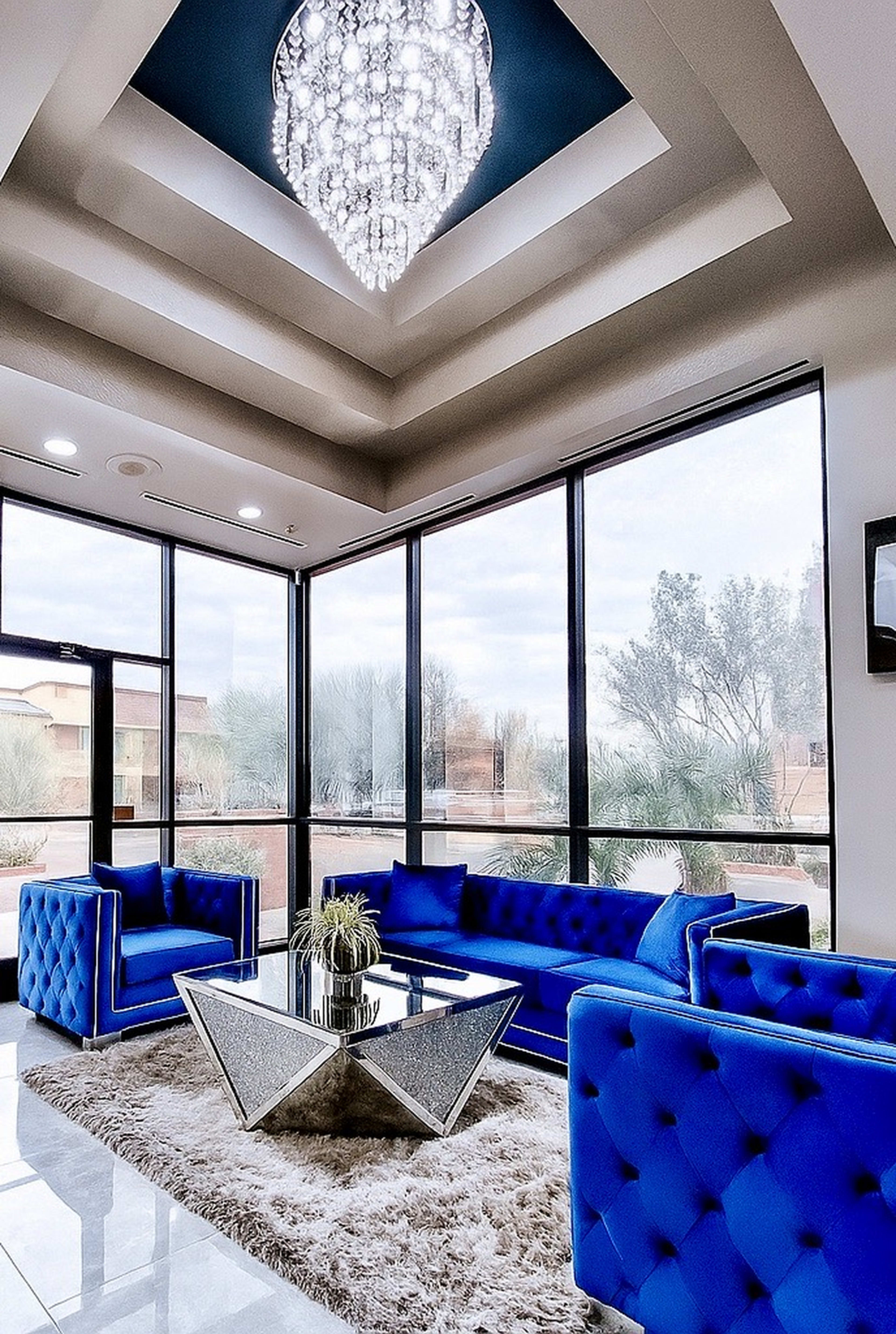 A living room features blue tufted sofas and chairs arranged around a mirrored coffee table, with a chandelier hanging from the ceiling and large windows letting in natural light.