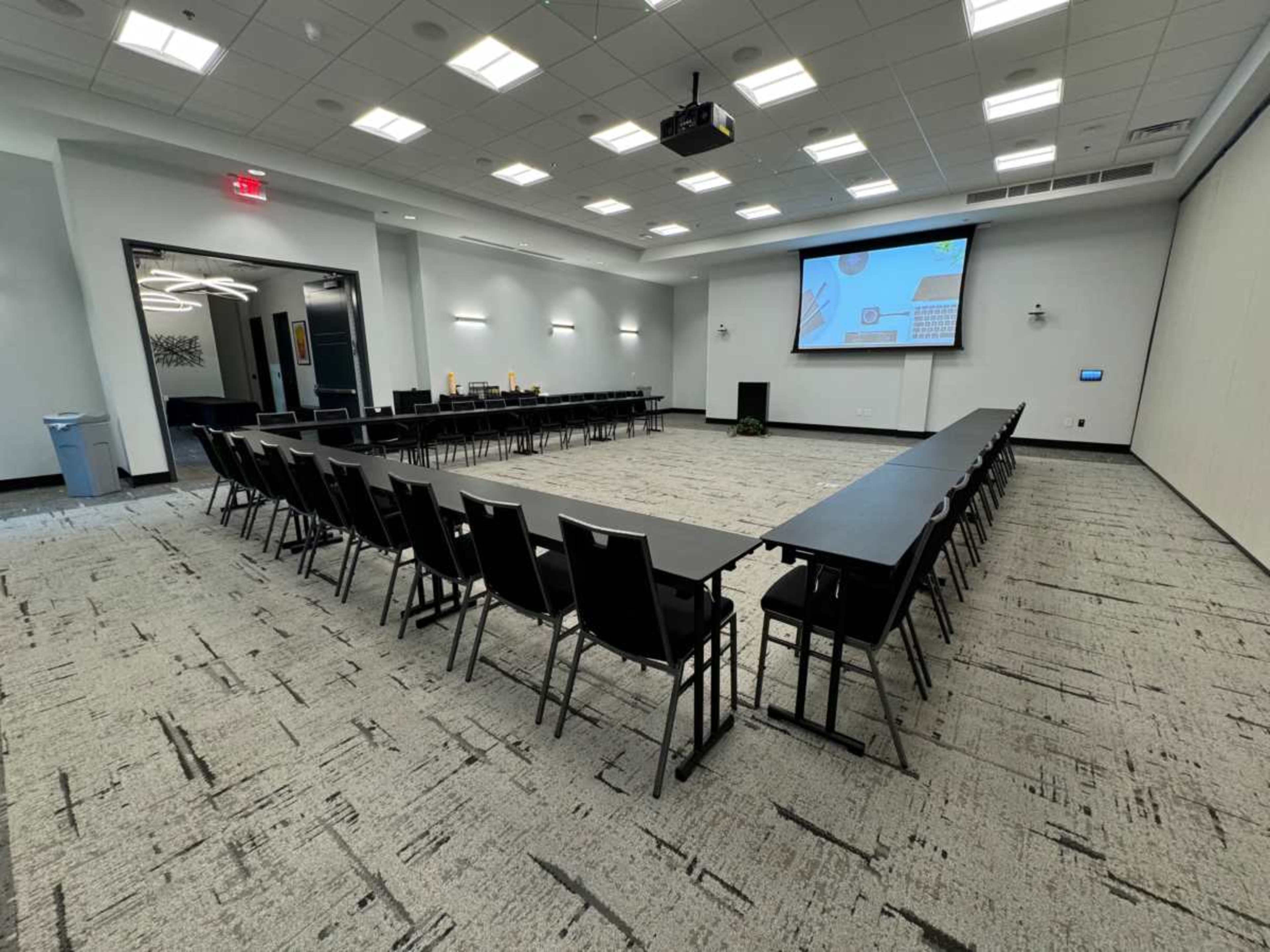 The image shows a spacious meeting room arranged with several rows of tables and chairs, featuring a large screen at one end and bright overhead lighting.