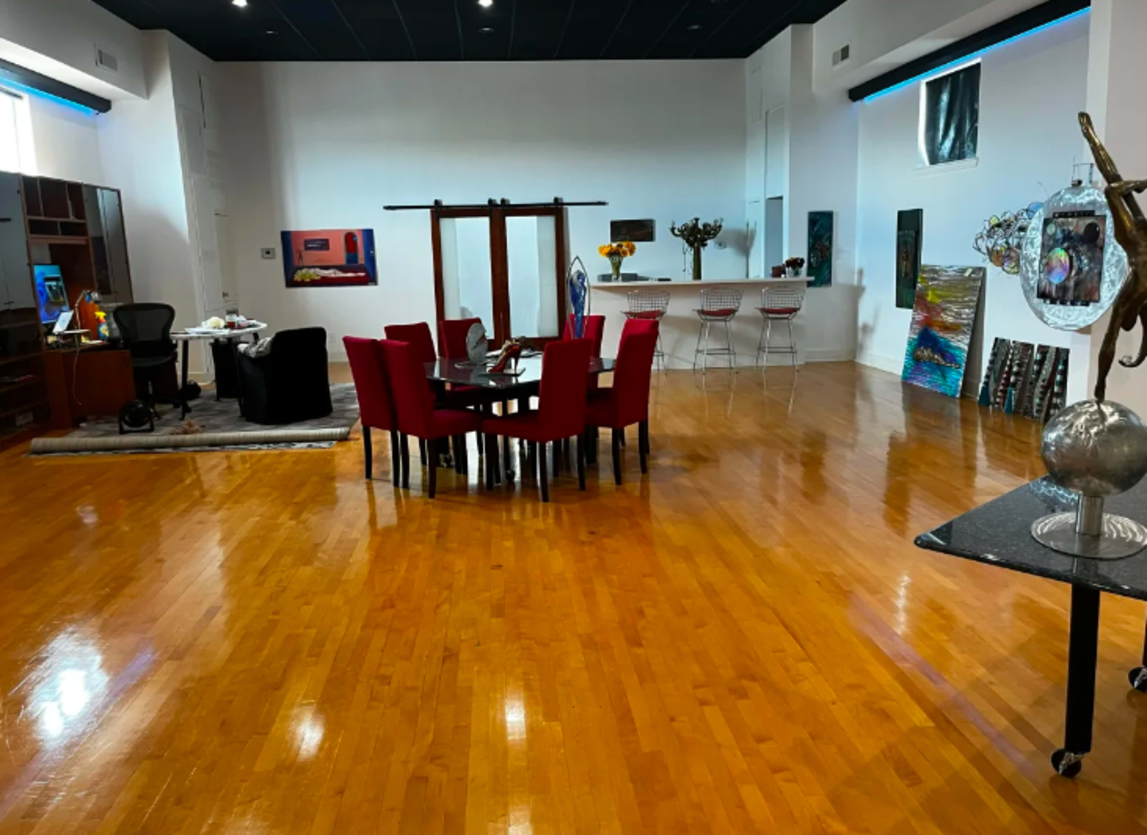 A spacious room with hardwood floors, featuring a round dining table surrounded by red chairs, along with various artworks displayed on the walls.