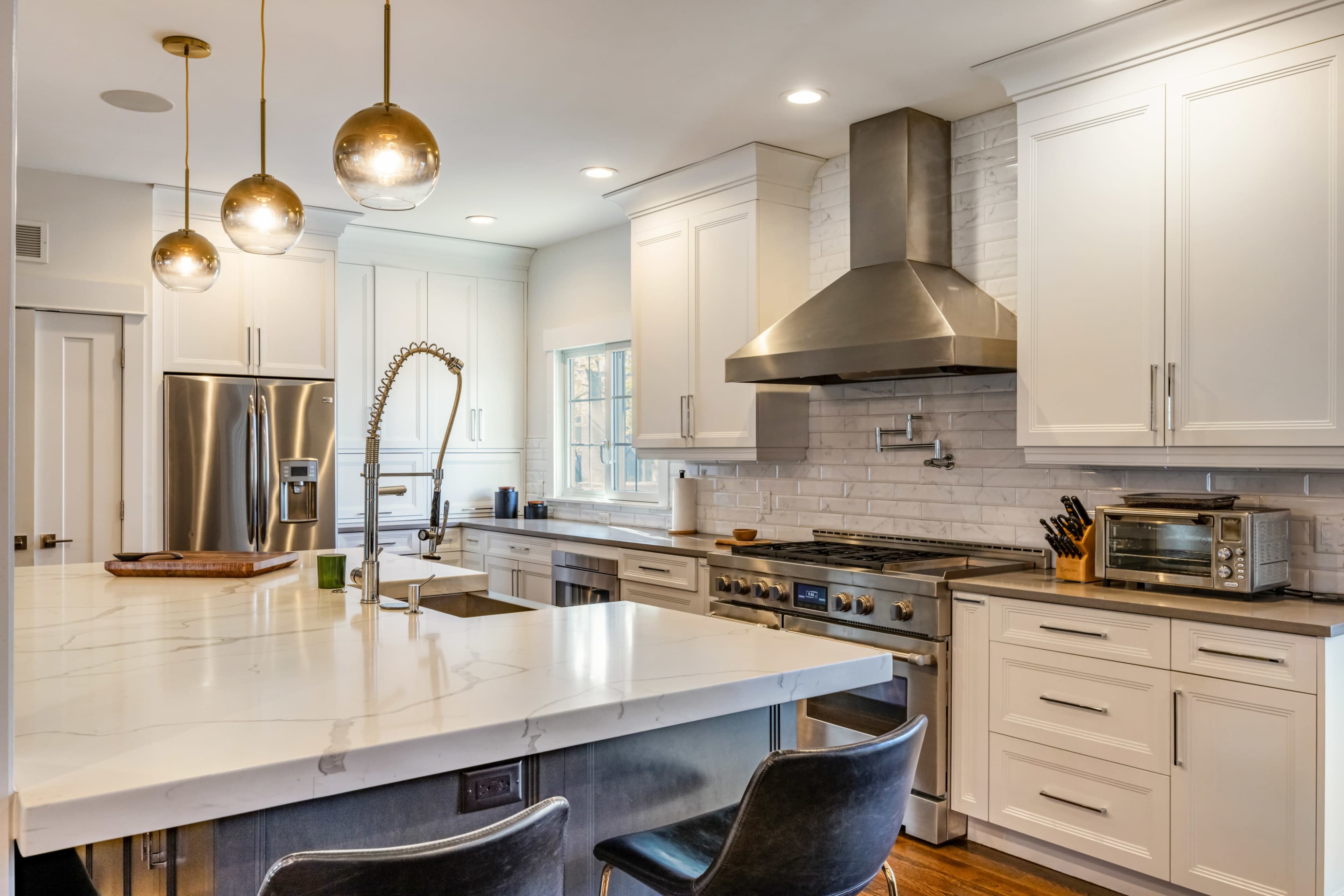 A modern kitchen features white cabinetry, stainless steel appliances, a large central island with a marble countertop, and stylish pendant lighting above.