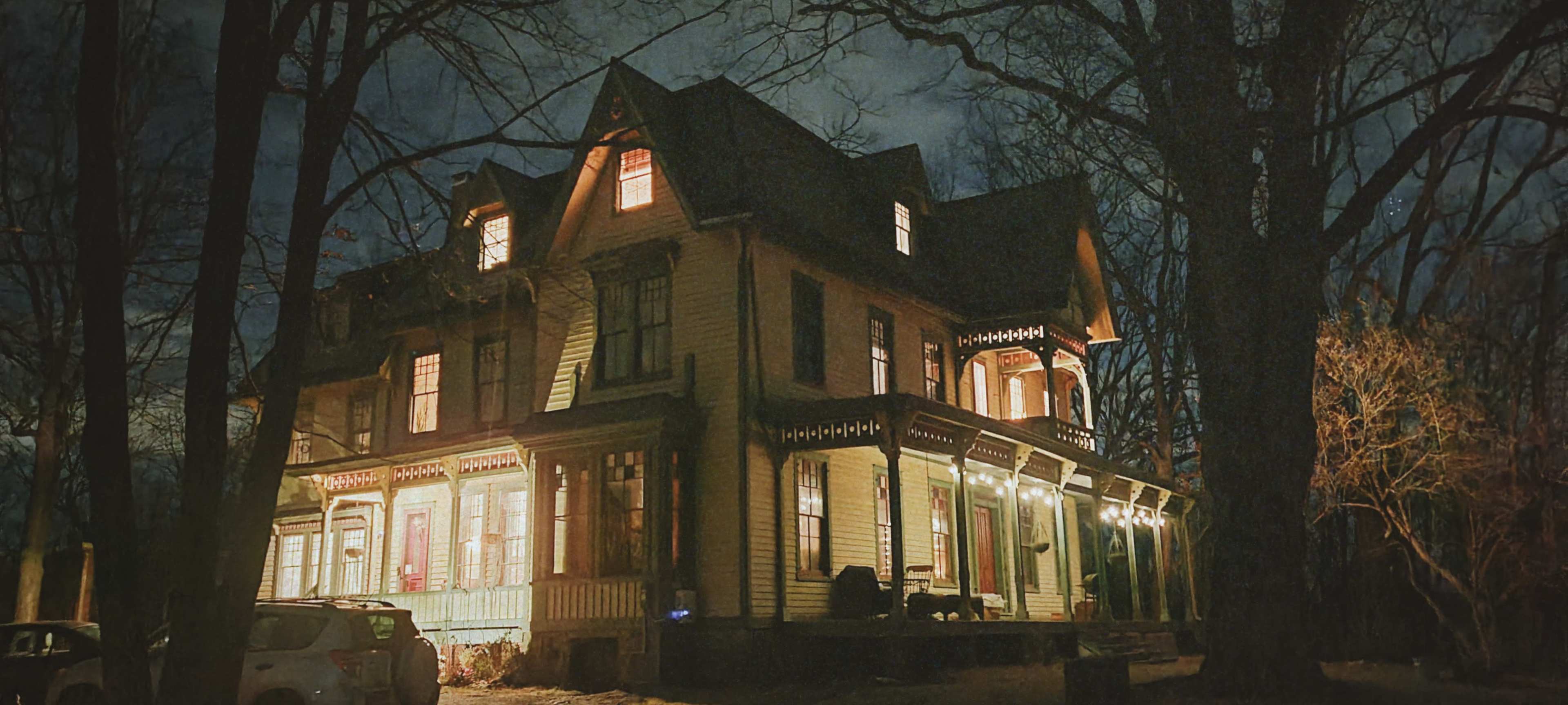 A large, gothic-style house is illuminated at night, showcasing its intricate architectural details and surrounded by bare trees.