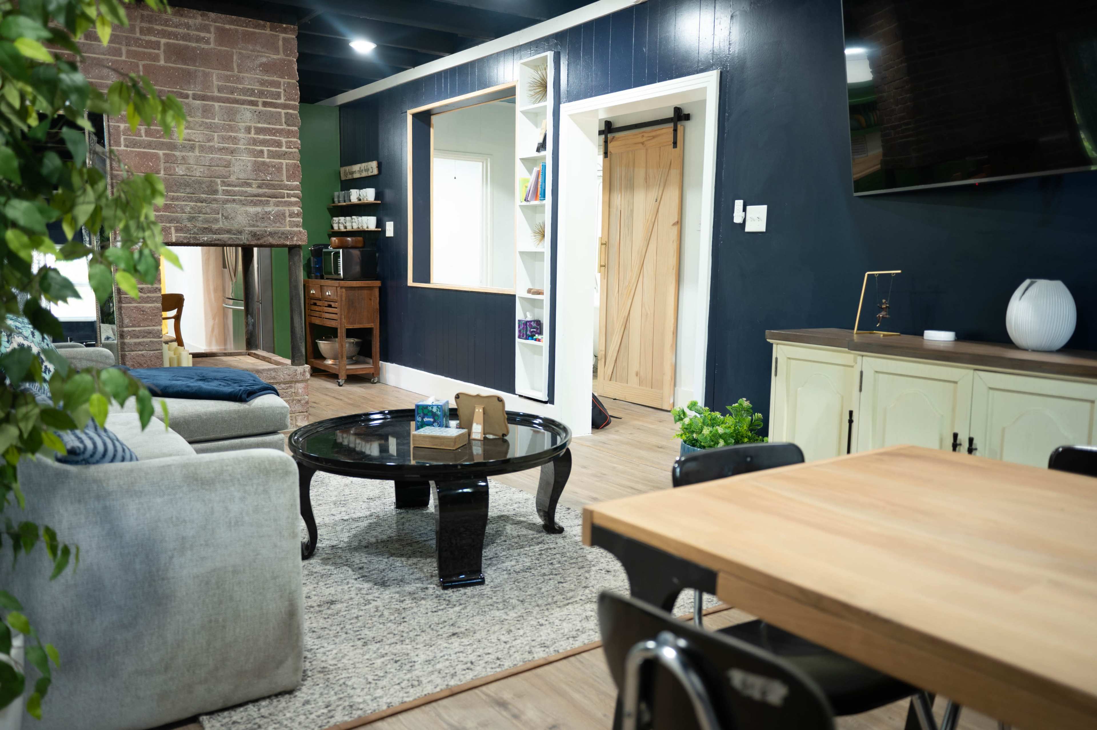 The image shows a modern living space featuring a sofa, a round coffee table, and a dining area with a wooden table, accompanied by green accents and a fireplace.