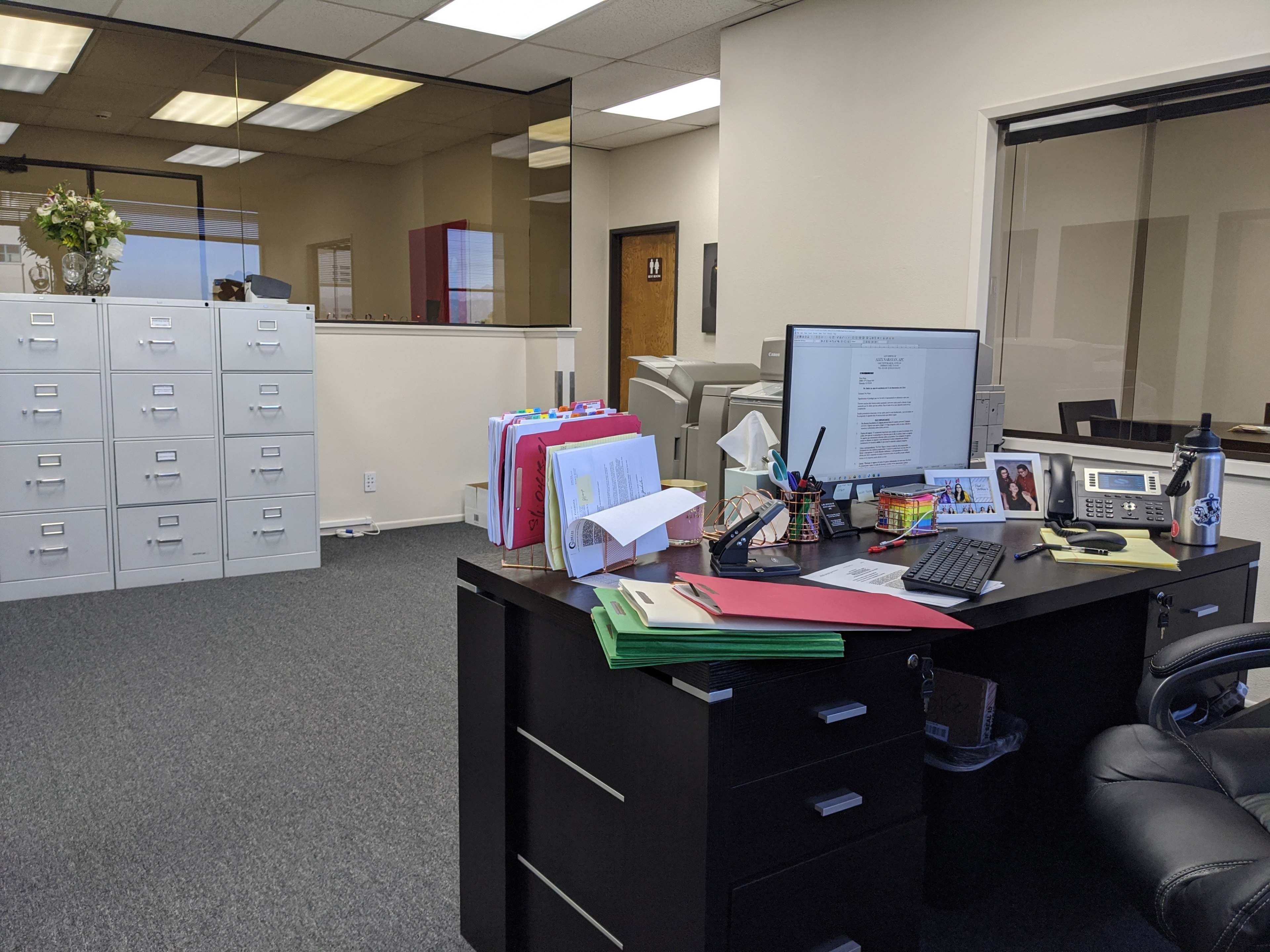 An office space with a black desk, a computer monitor, and colorful file folders, along with filing cabinets visible in the background.