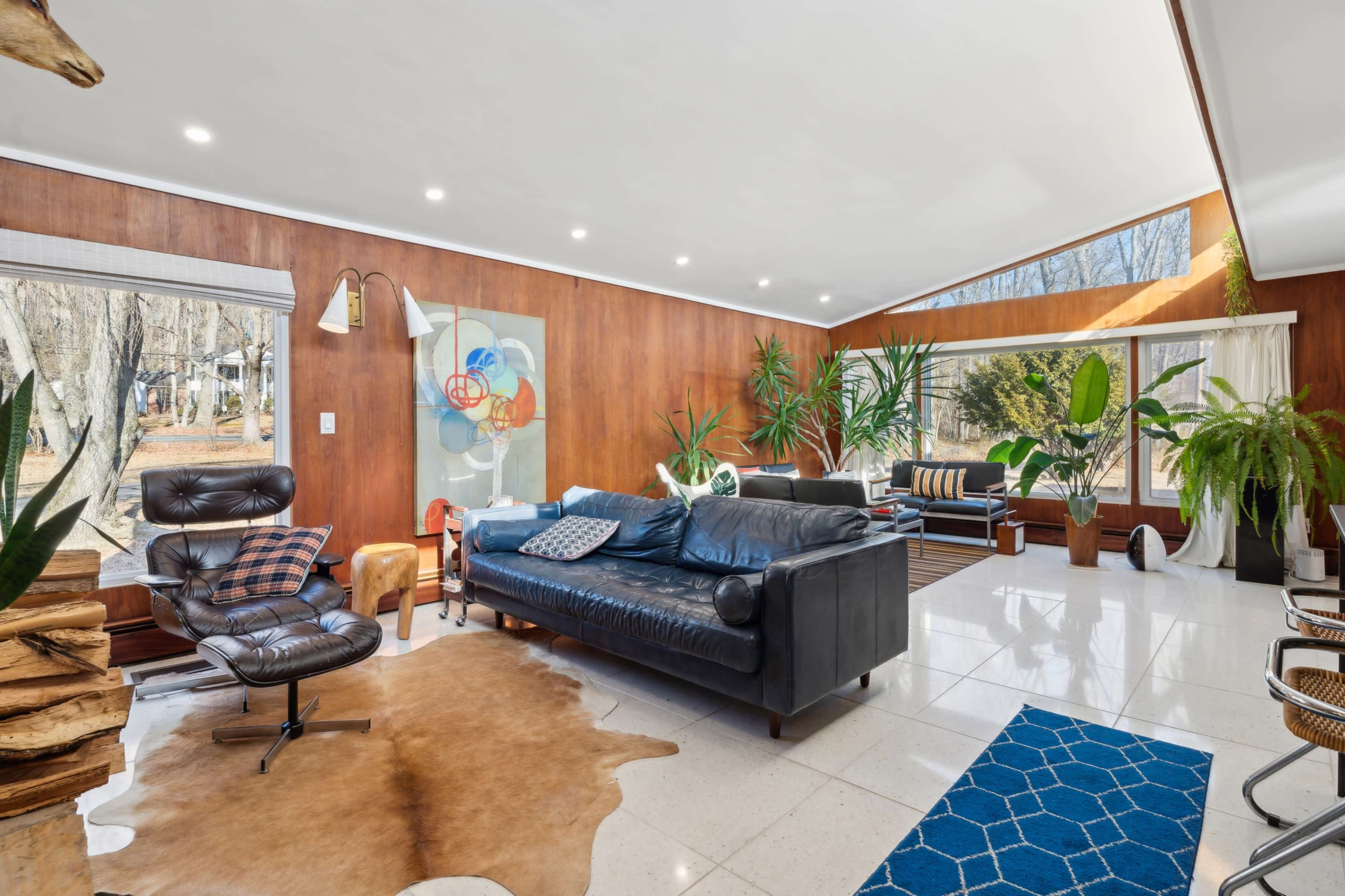 The image shows a modern living room with wooden walls, a blue leather couch, and large windows allowing natural light to fill the space, along with decorative plants.