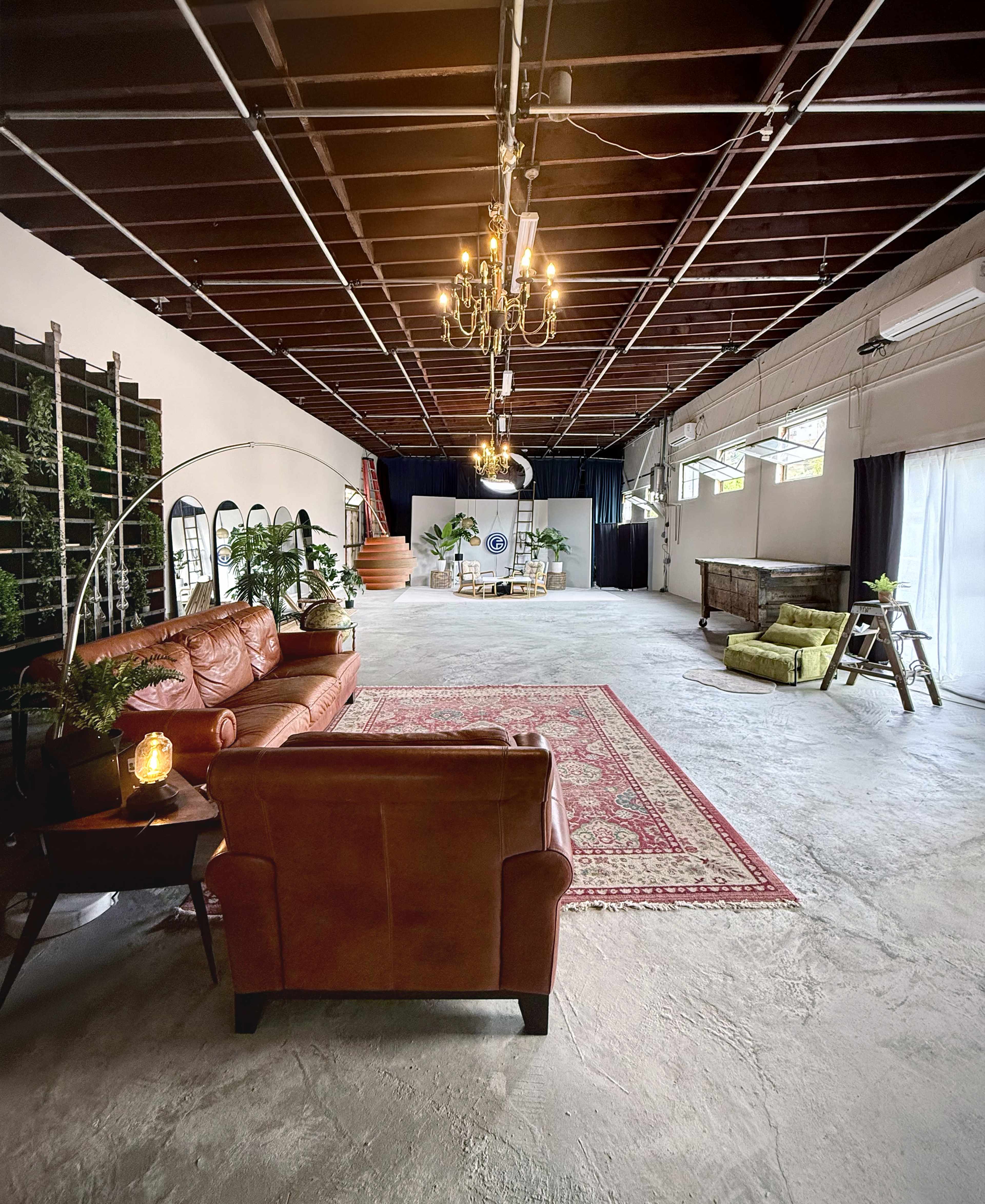 The image shows a spacious industrial-style room with a mix of seating areas, including a brown leather sofa and a green chair, along with decorative plants and a rug on a concrete floor.