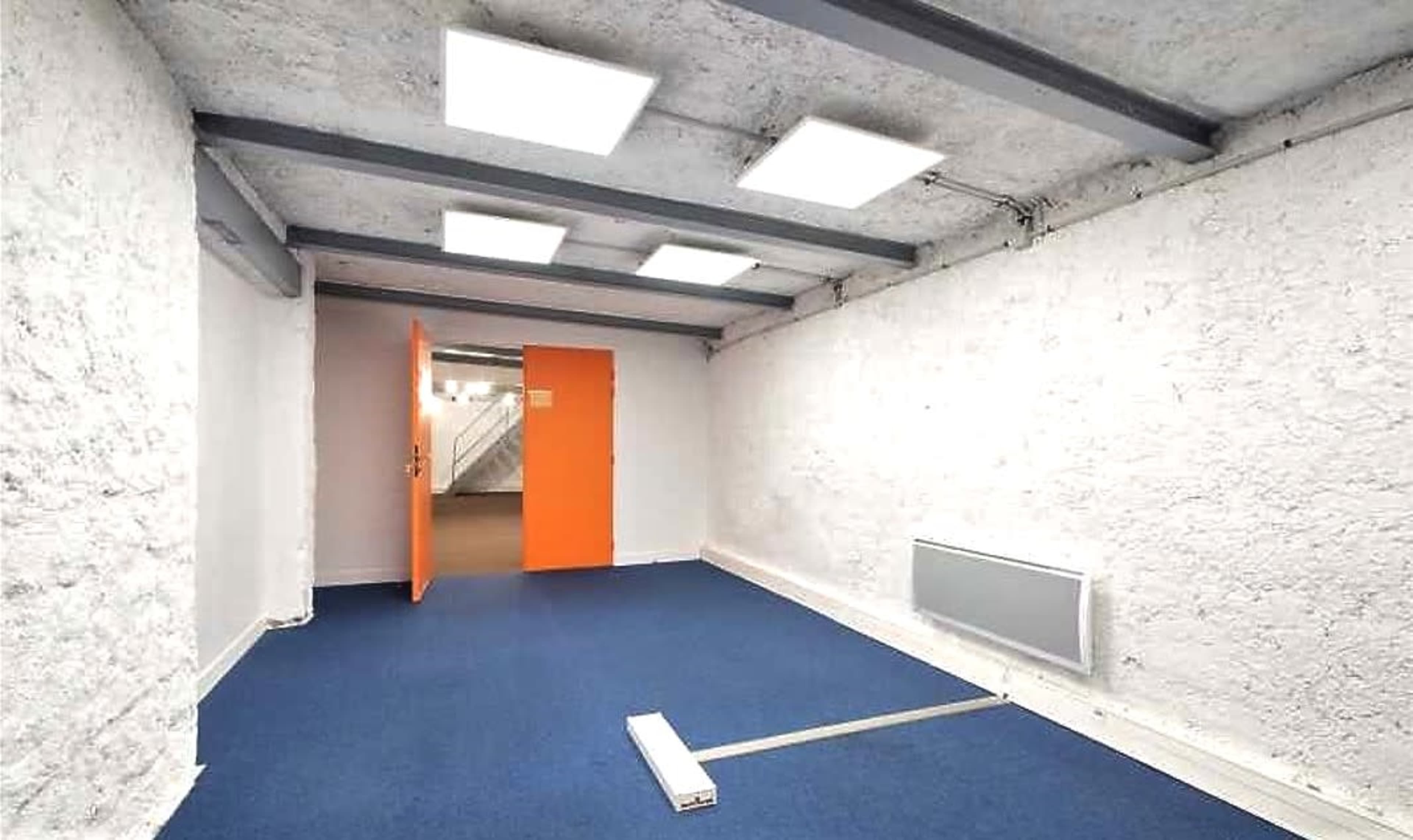 A sparsely furnished room with a blue carpet, white walls, and an orange door leading to another space.