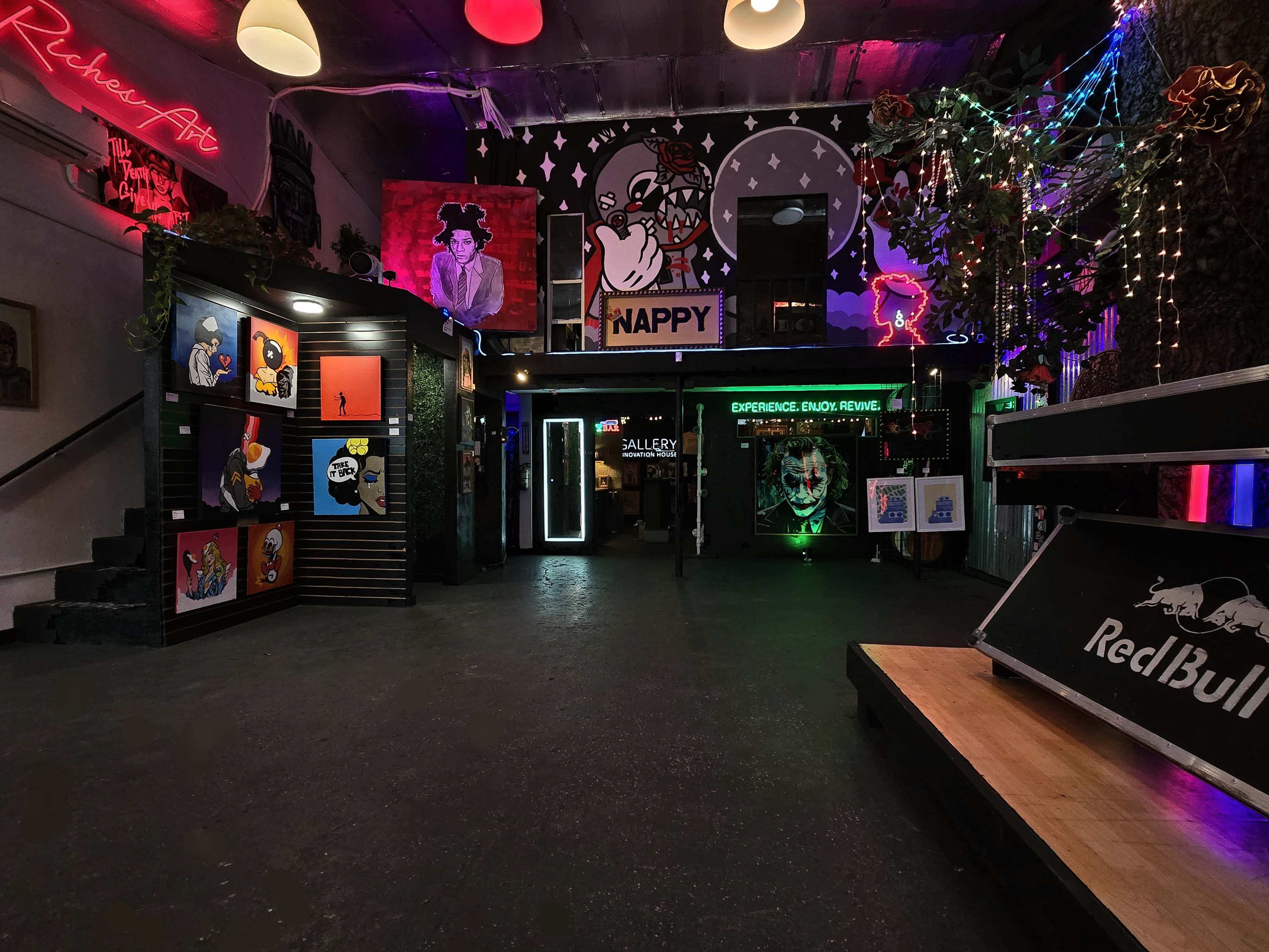 The image shows a vibrant indoor space featuring colorful wall art, neon lights, and various installations, including a stage area and a gallery display.