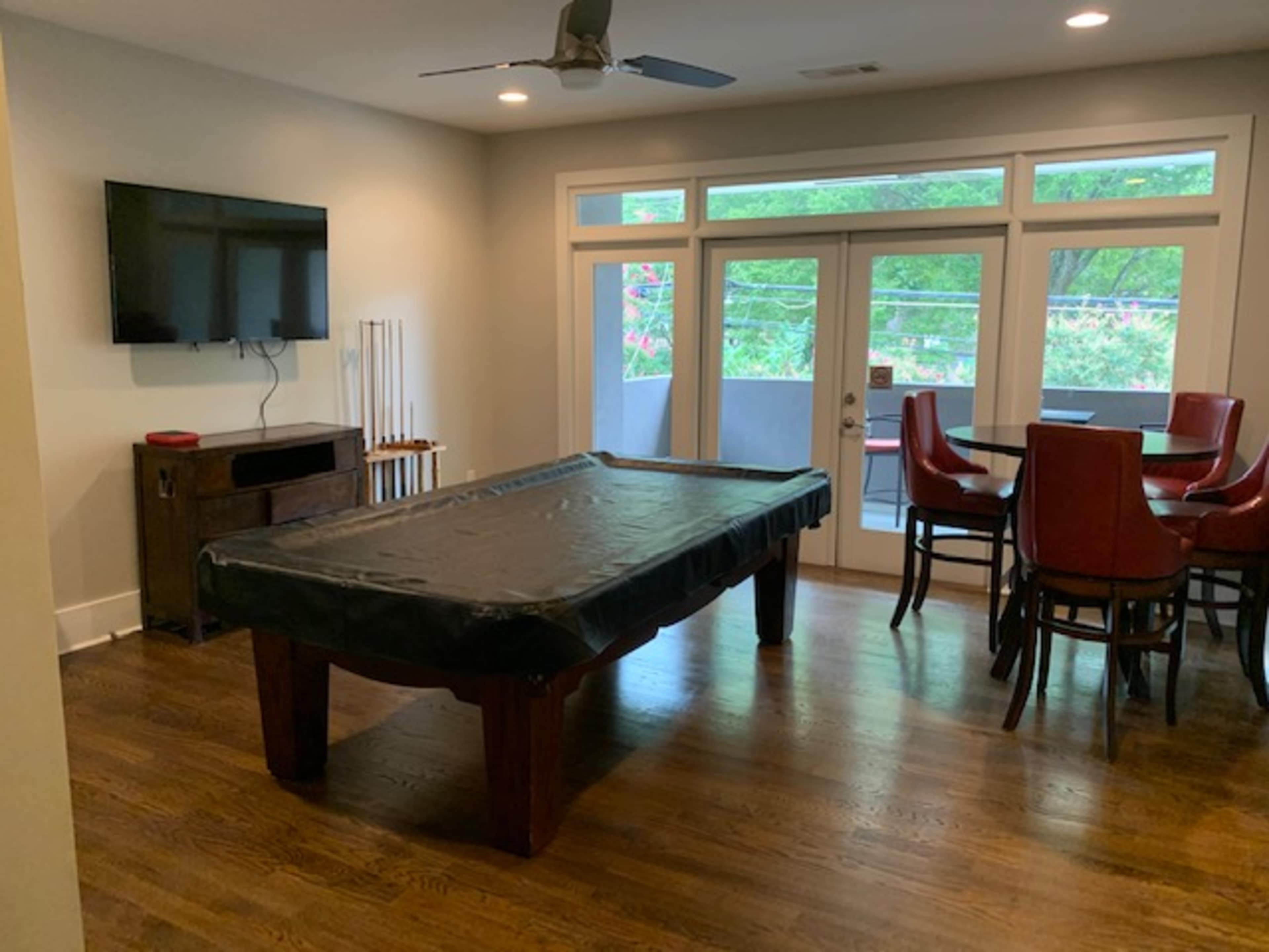 A room containing a covered pool table, a wall-mounted television, and a dining area with red chairs near large windows.