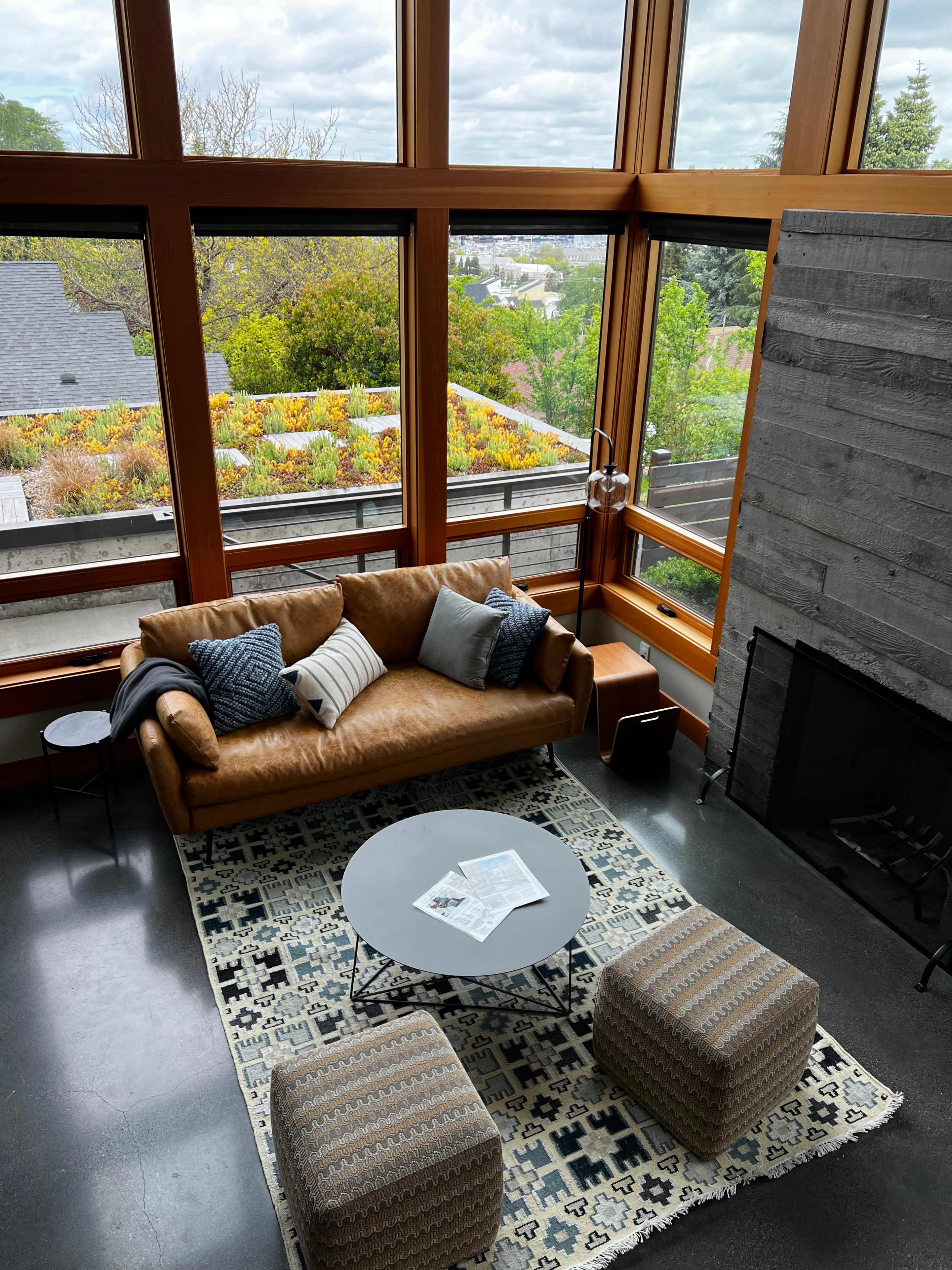 The image shows a modern living room with a large leather sofa, a round coffee table, and a textured area rug, featuring large windows that overlook a green landscape.