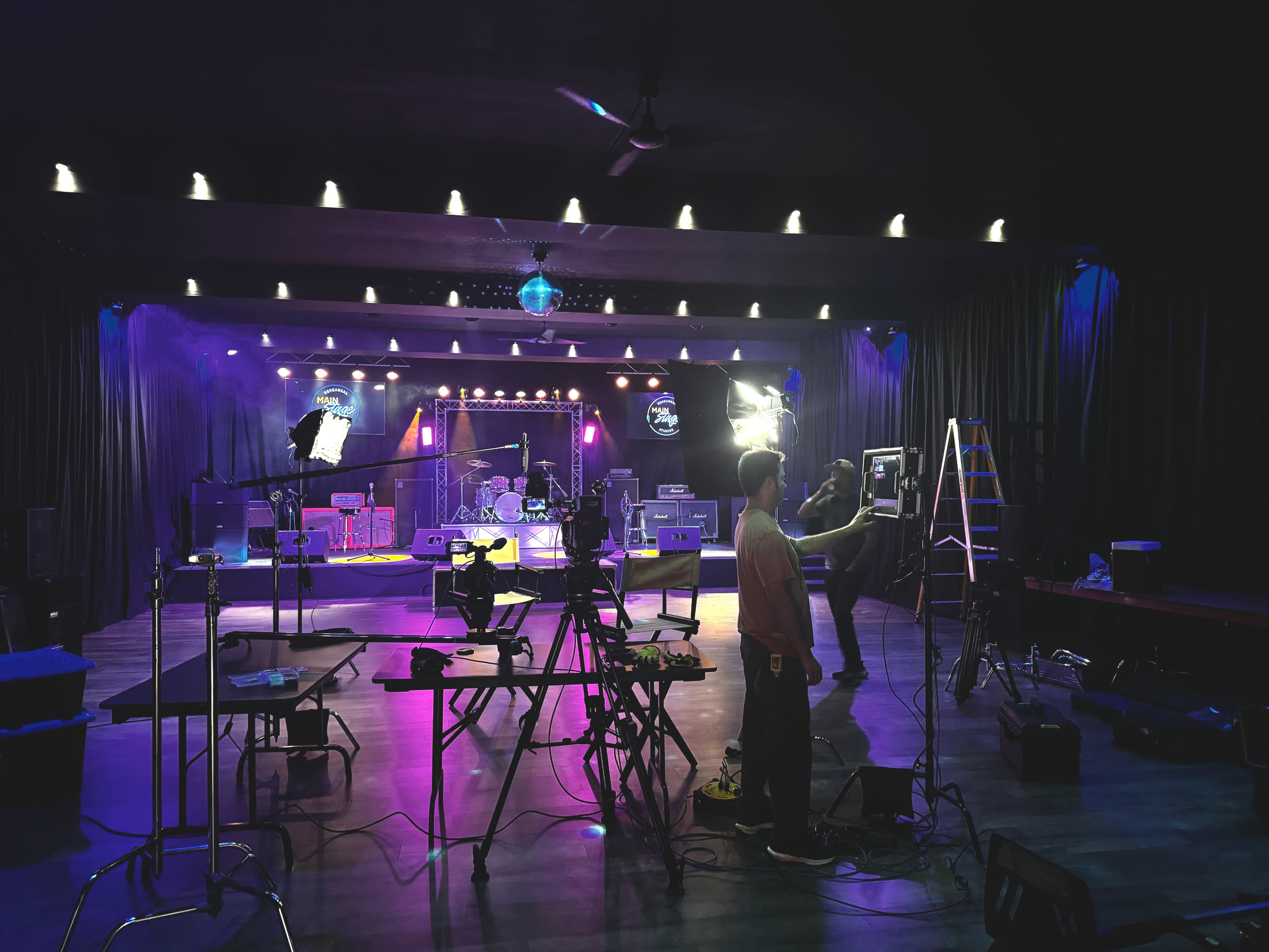 A dimly lit performance venue is set up for a concert, featuring musical equipment, lighting rigs, and a small stage with instruments in the background.