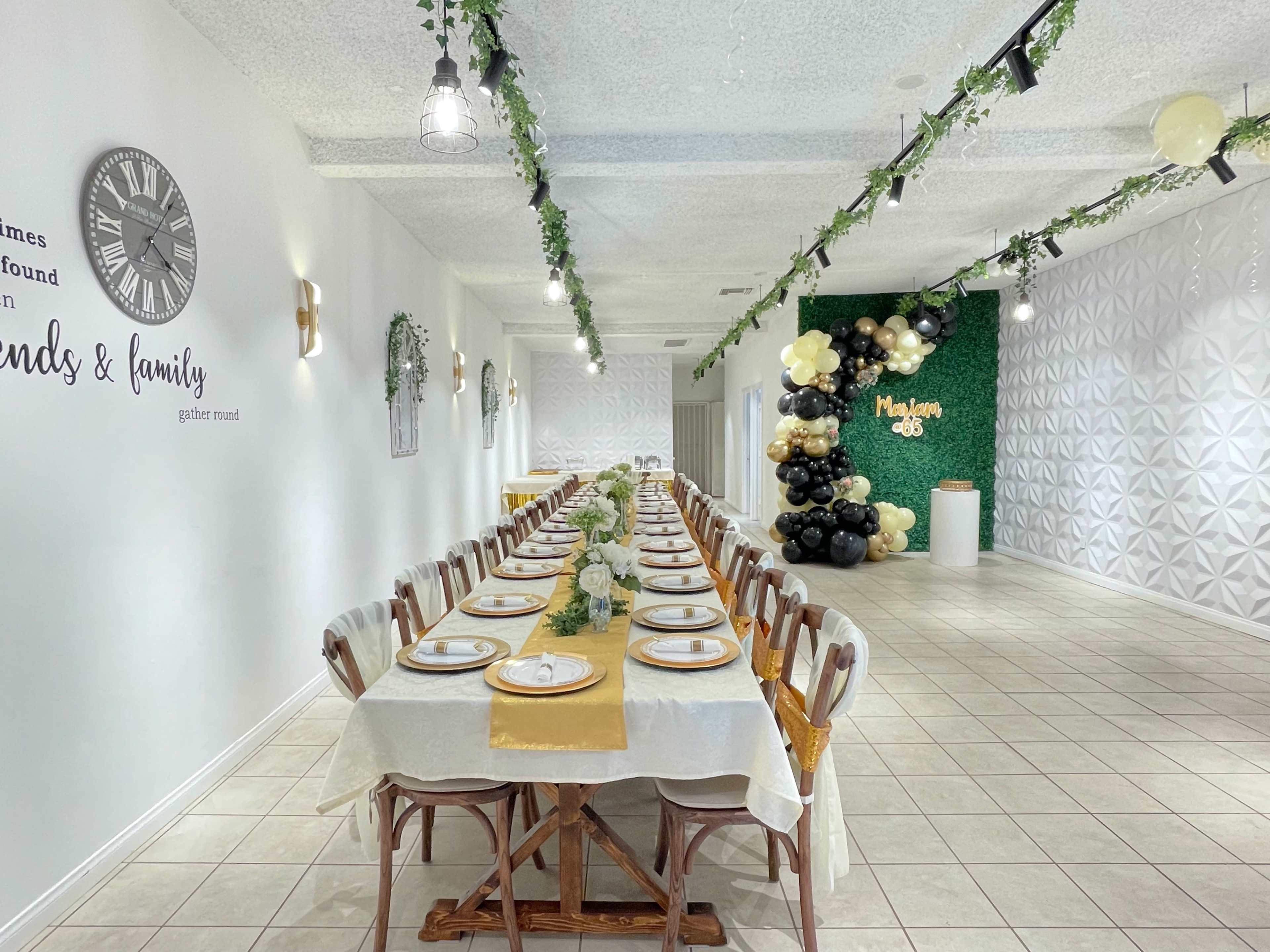 A long banquet table is set with elegant white tablecloths and gold accents in a decorated dining space featuring greenery and a large balloon arrangement.