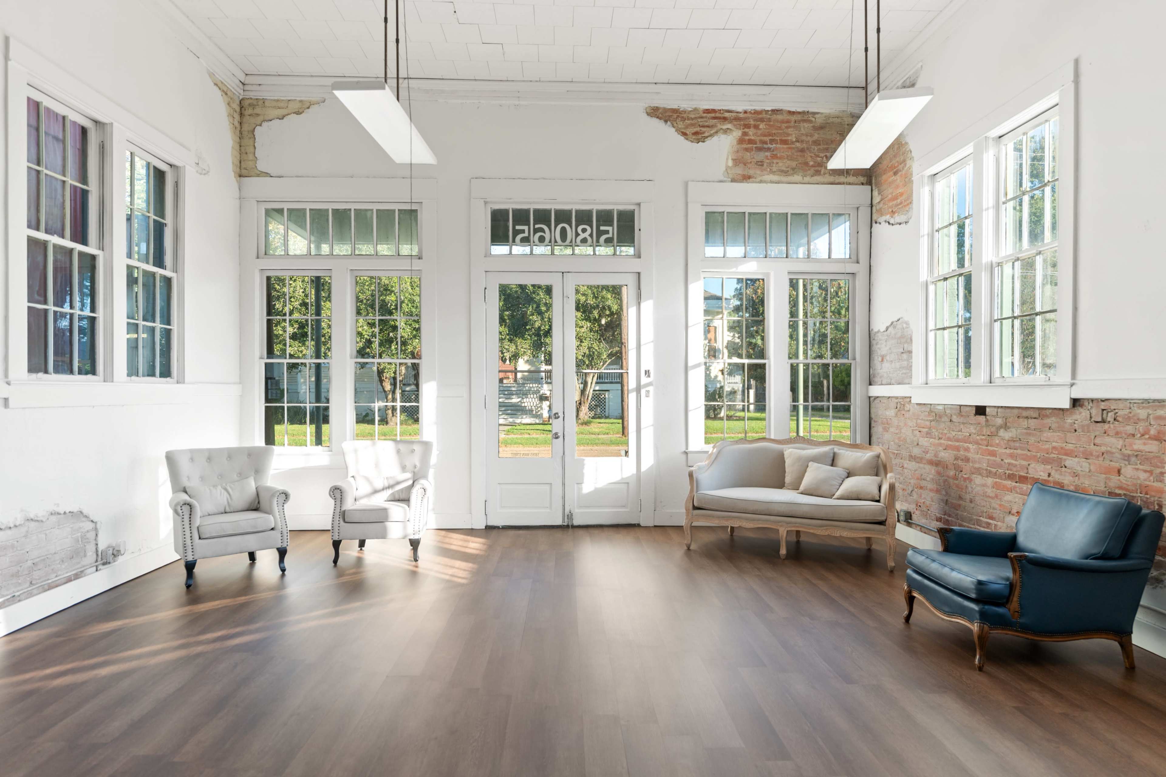 The image shows a spacious room with large windows, featuring a light wooden floor and several seating arrangements including armchairs and a couch.