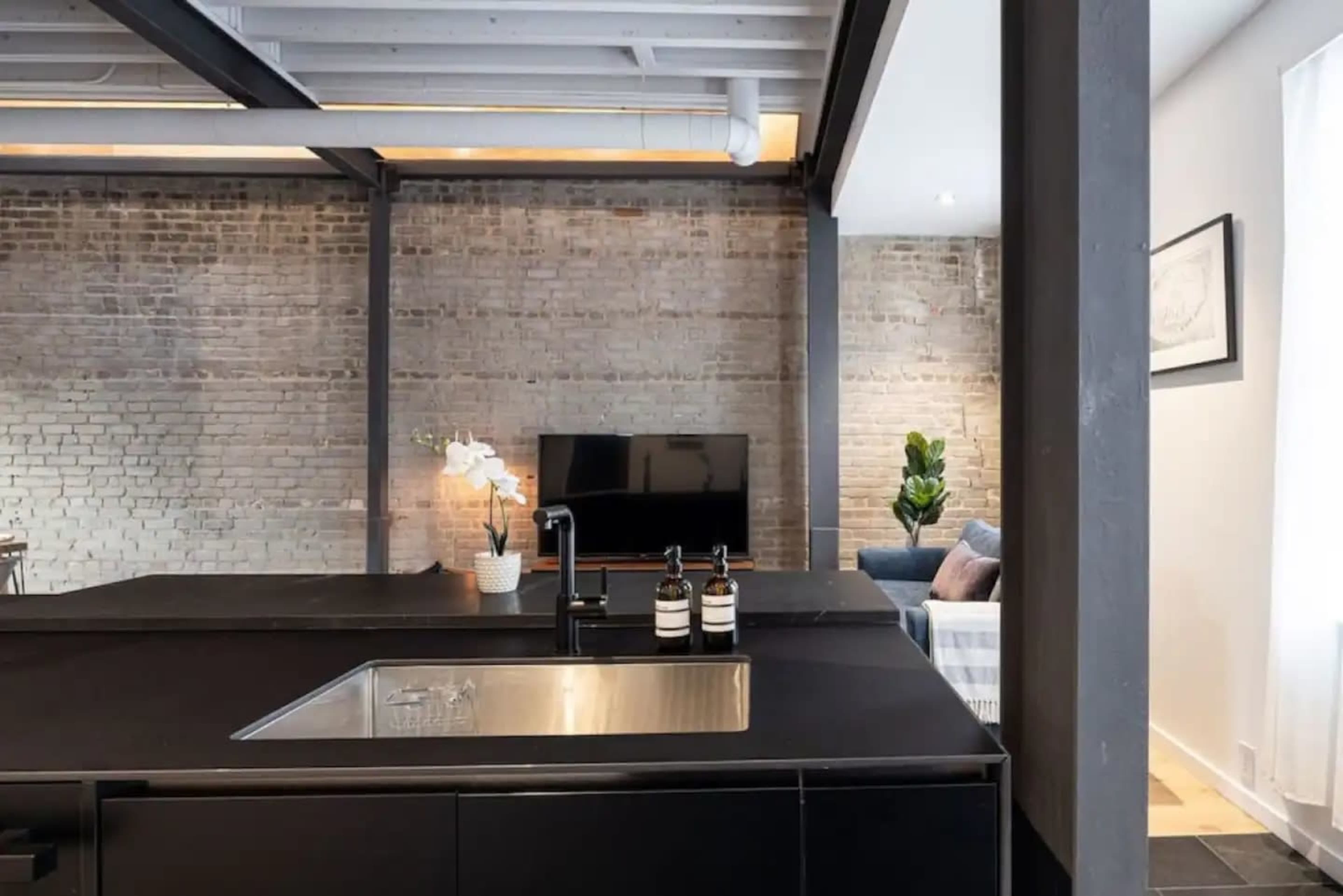 A modern kitchen features a black countertop and sink, with a view of a living area that includes a TV and a couch against a brick wall.