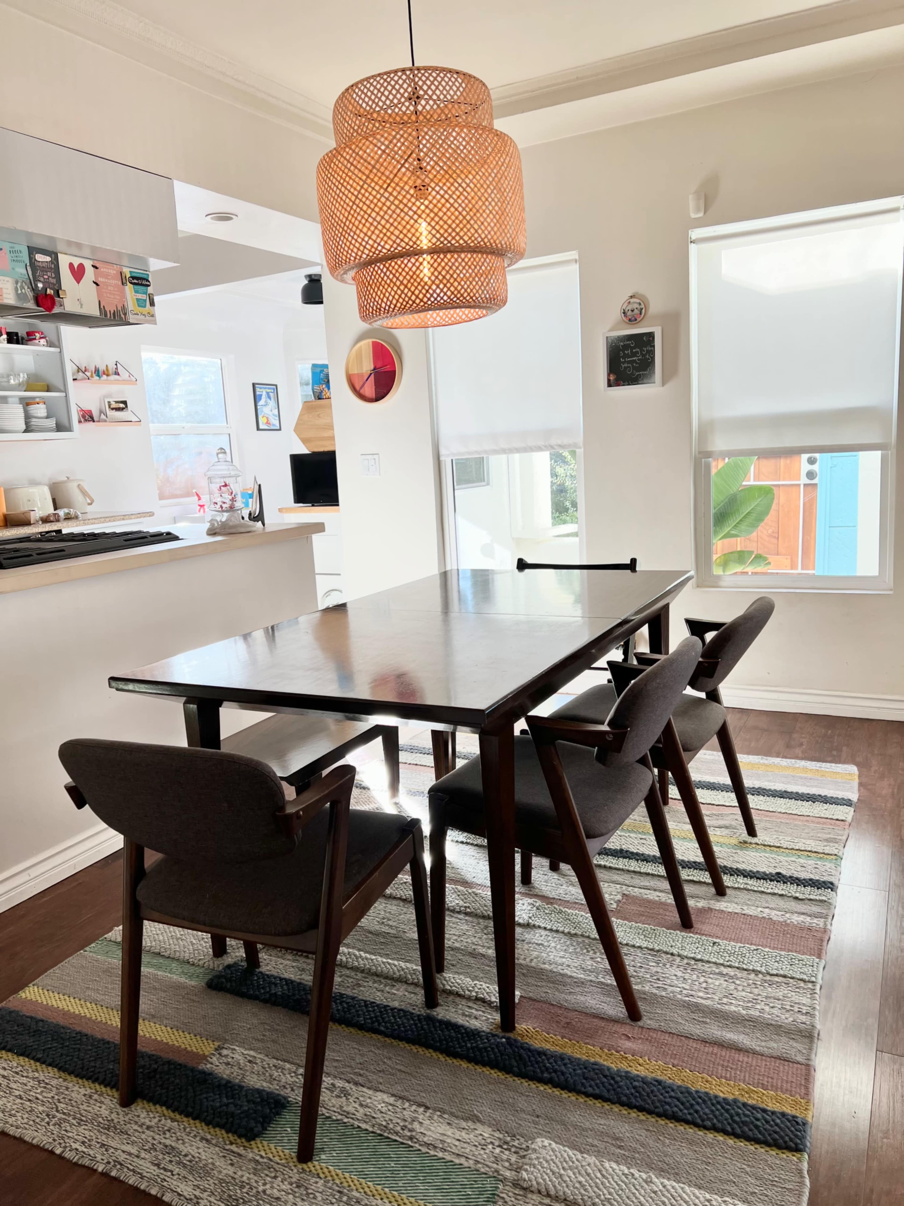 A dining area features a wooden table surrounded by four chairs, a woven pendant light, and a striped rug on a hardwood floor.