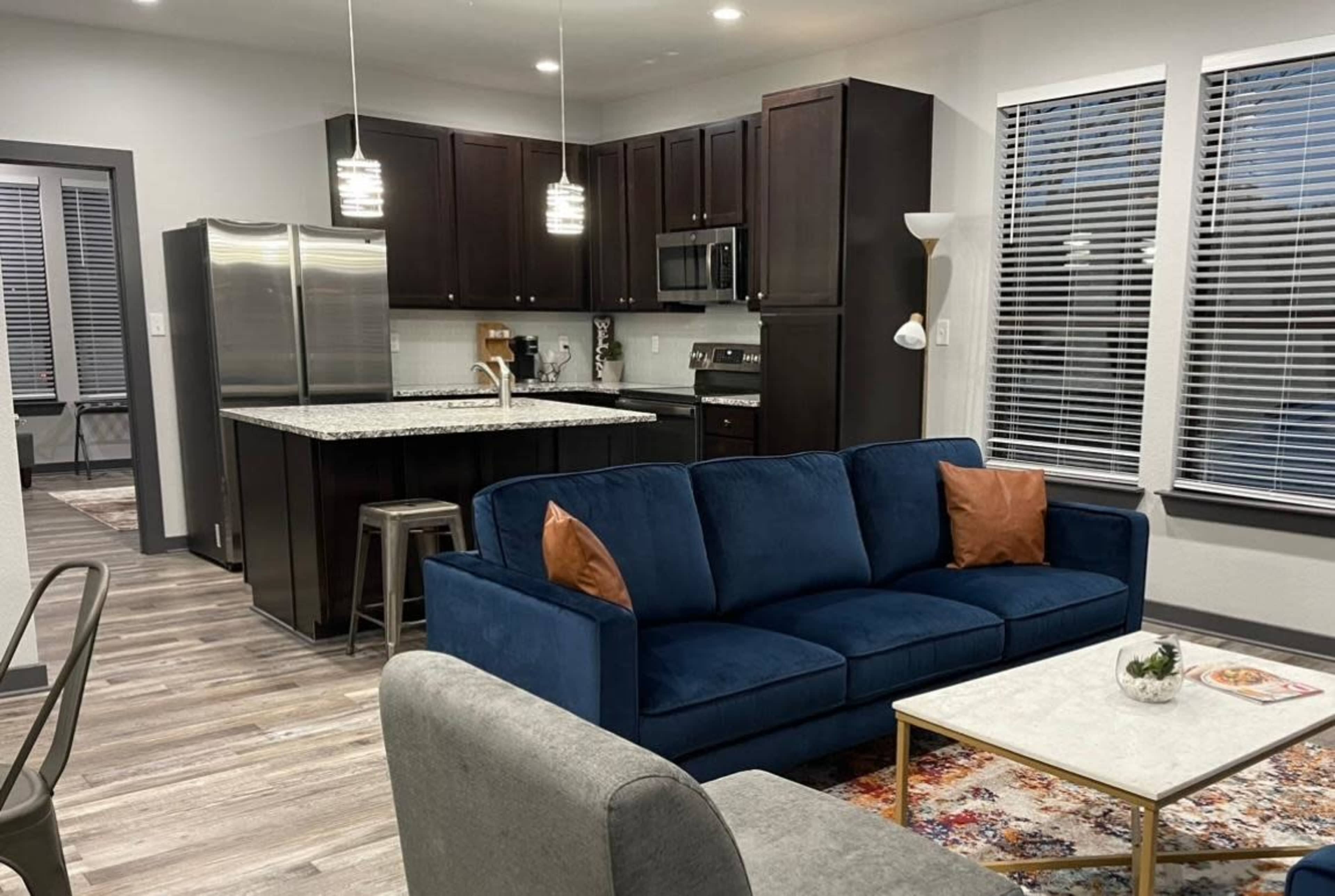 A modern living room with a blue sofa, a light-colored coffee table, and an open kitchen featuring dark cabinetry and stainless steel appliances.