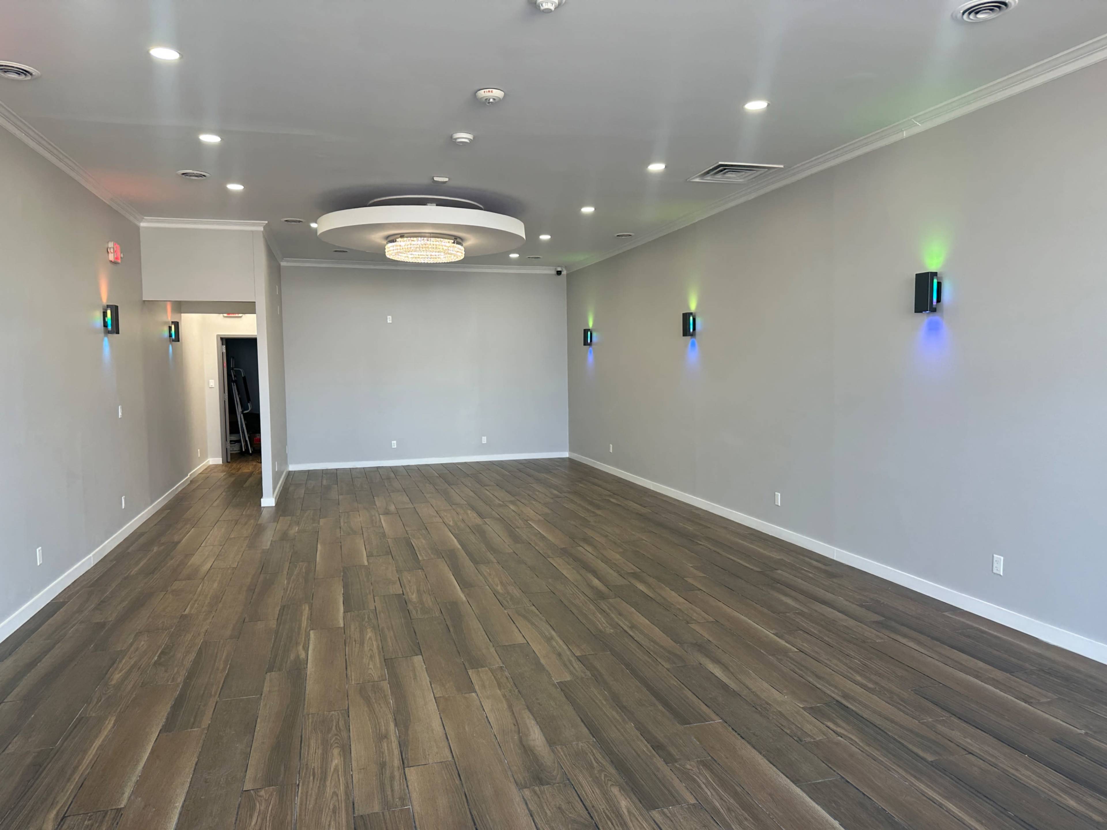 The image shows a spacious, modern room with wooden flooring, white walls, and decorative lighting fixtures on the walls.
