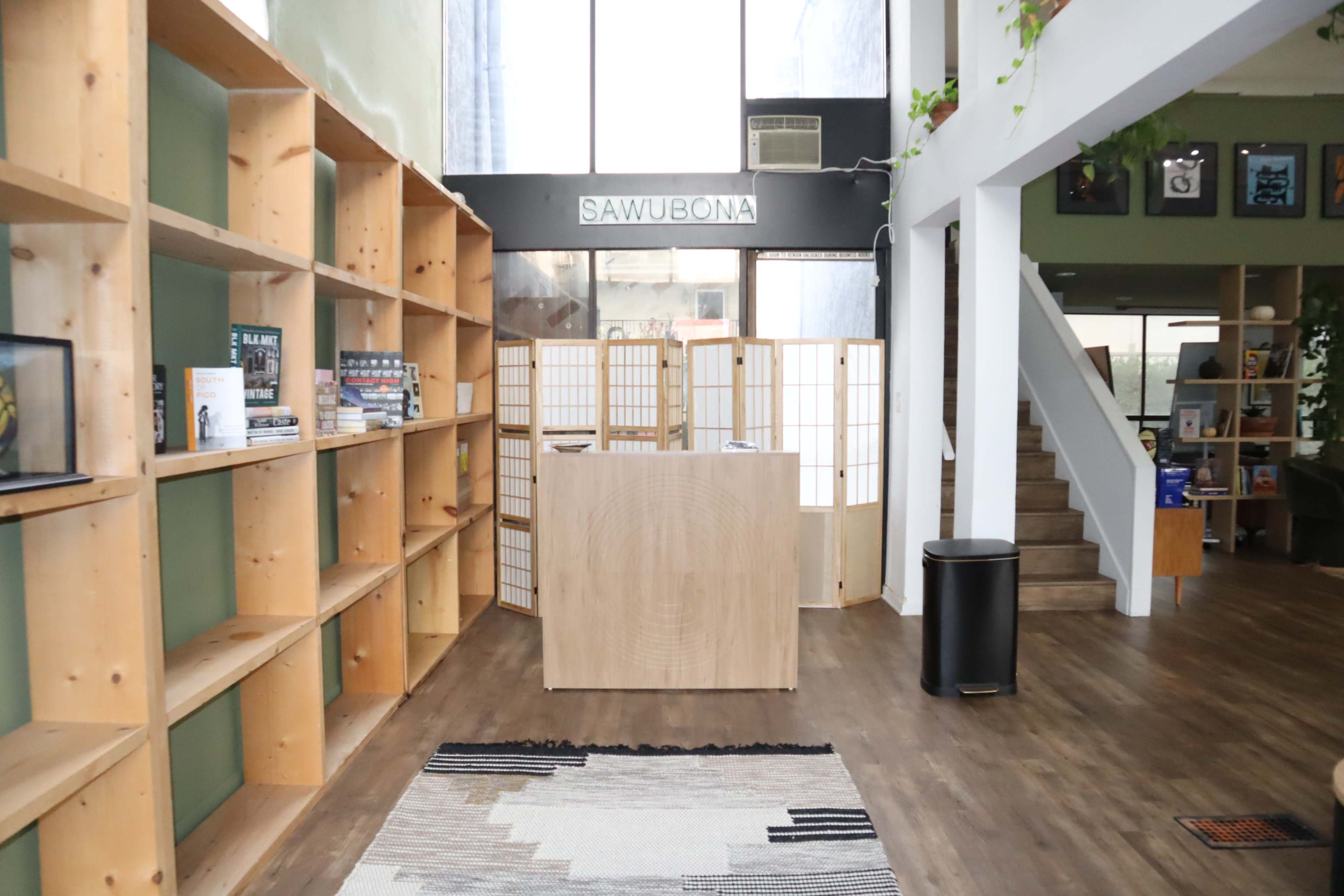 The image shows a spacious interior with wooden shelving units filled with books, a reception desk in the center, and a staircase leading to an upper level.