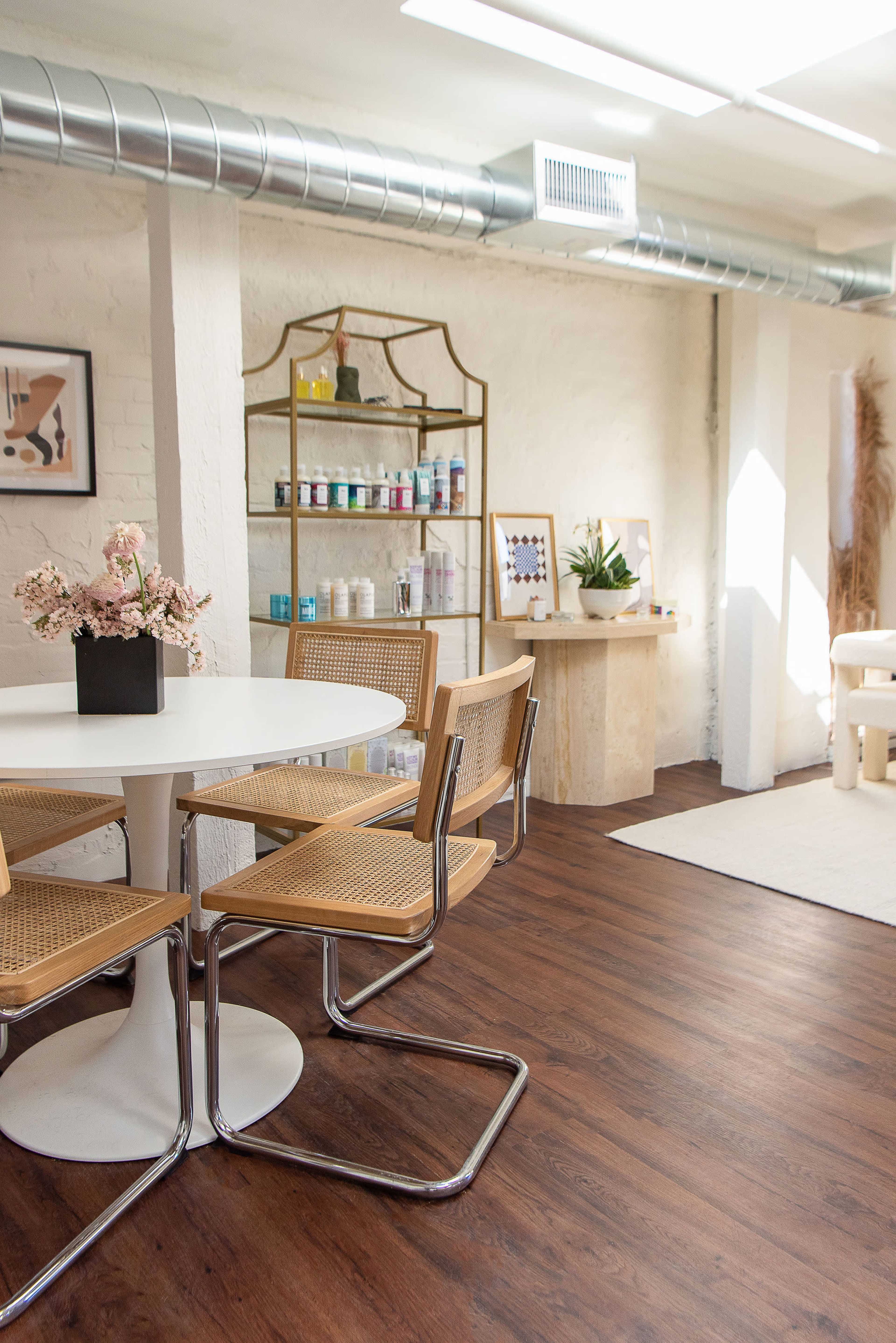 A bright, modern interior space with a round white table surrounded by four woven chairs, a shelf displaying various products, and a cozy seating area with plants and decor elements.