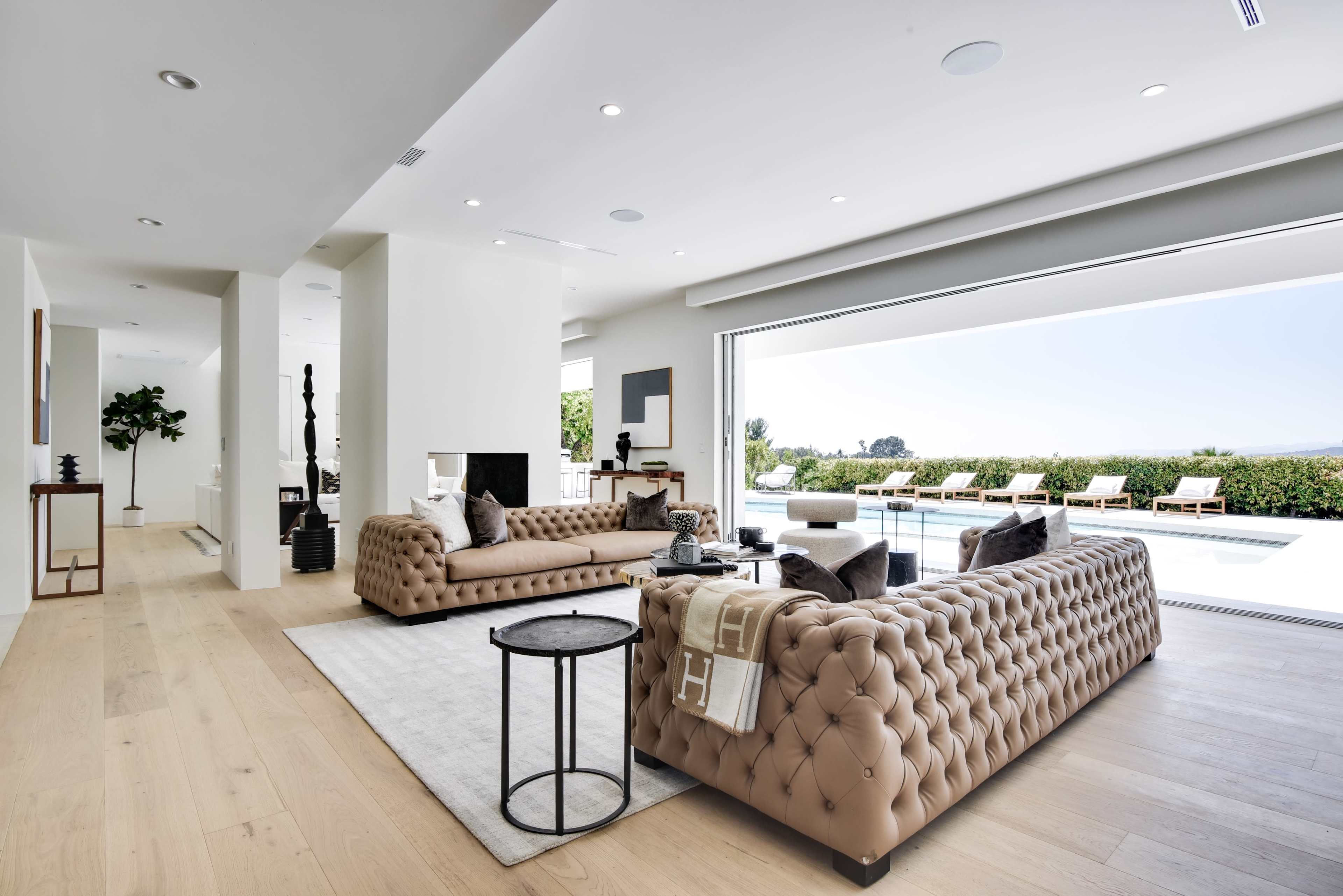The image shows a modern living room featuring two brown tufted sofas facing each other, with large glass doors opening to a pool area and landscaped outdoor space.