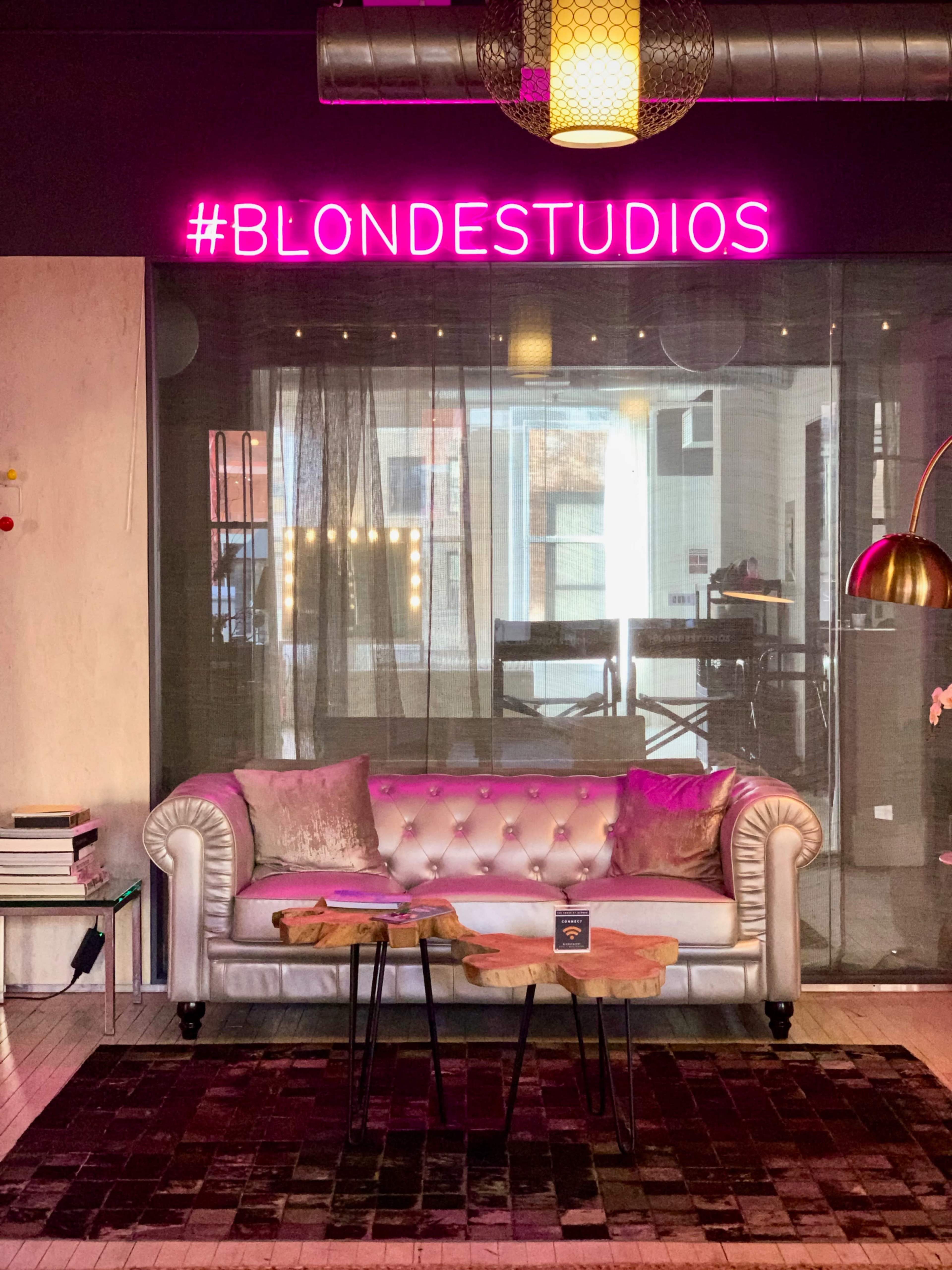The image shows a stylish interior featuring a tufted silver sofa, a neon pink hashtag sign reading "#BLONDESTUDIOS," and wooden tables in front of the sofa.