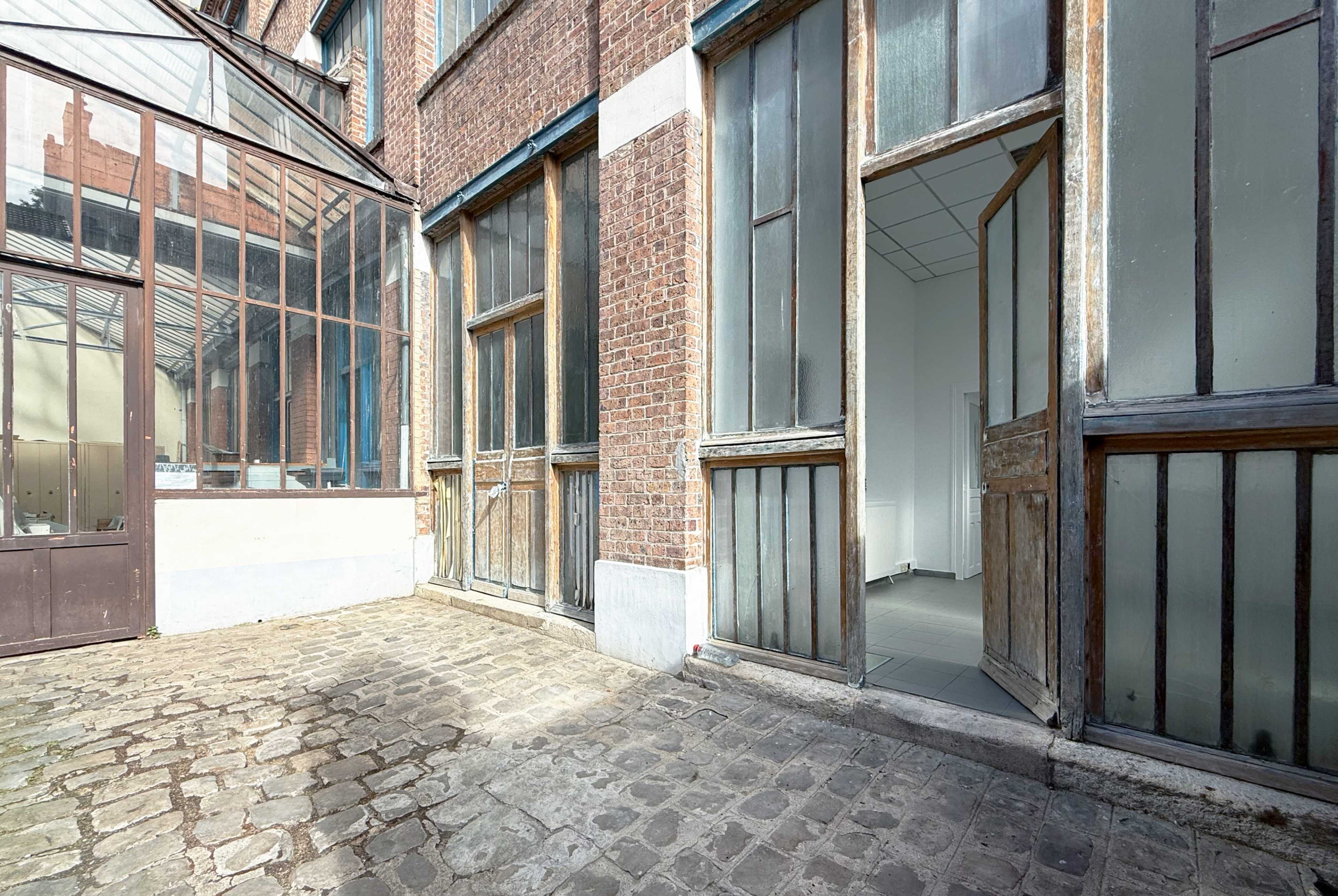 The image shows a cobblestone courtyard enclosed by brick walls and glass windows, featuring wooden doors that open into an interior space.