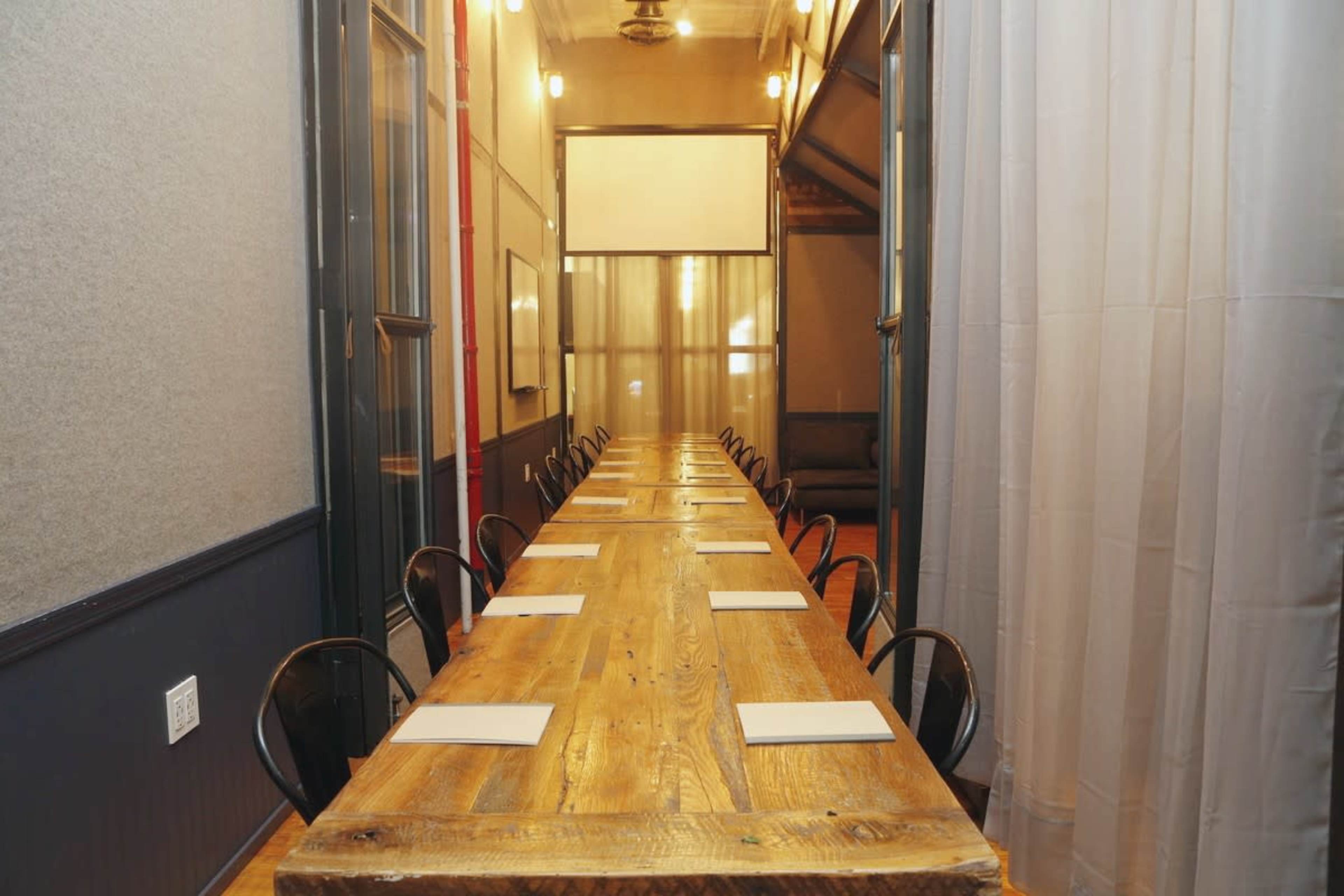 A long wooden table with chairs is set up in a conference room featuring large windows and a projector screen at the far end.