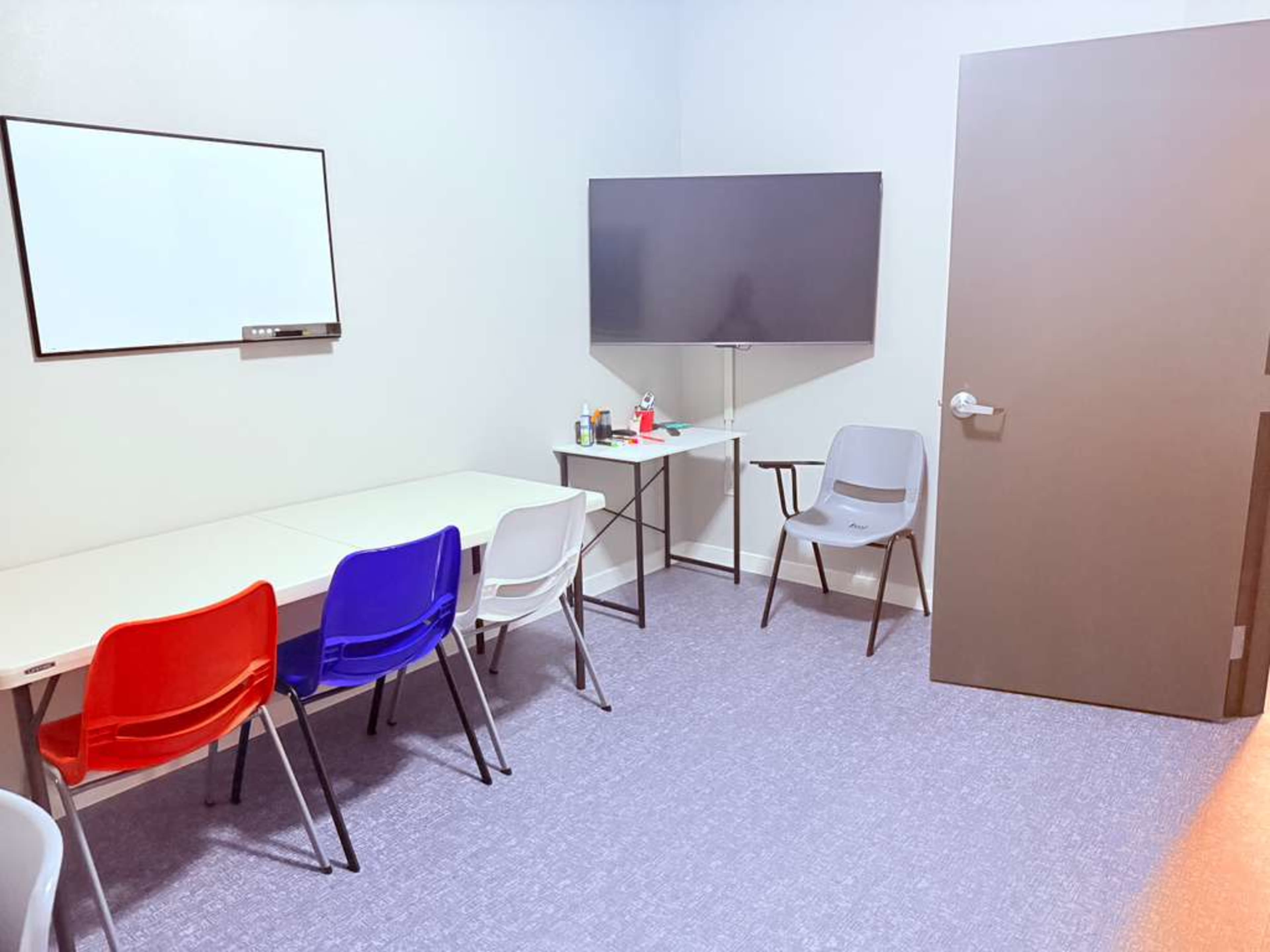 The image shows a small meeting room equipped with a whiteboard, a large television, a table, and several chairs in different colors.