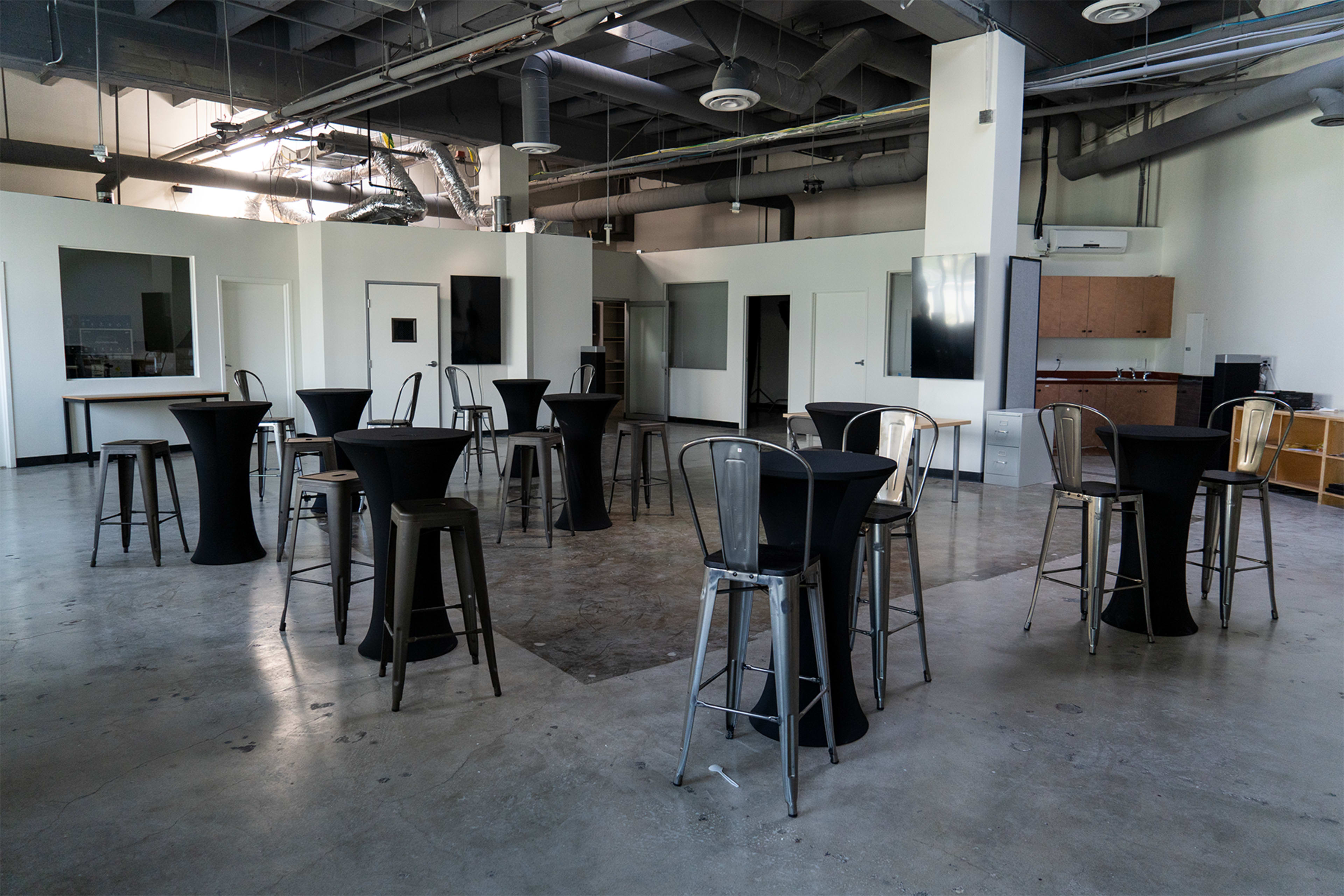 A spacious room with high tables and metal stools arranged in the center, surrounded by white walls and large windows.