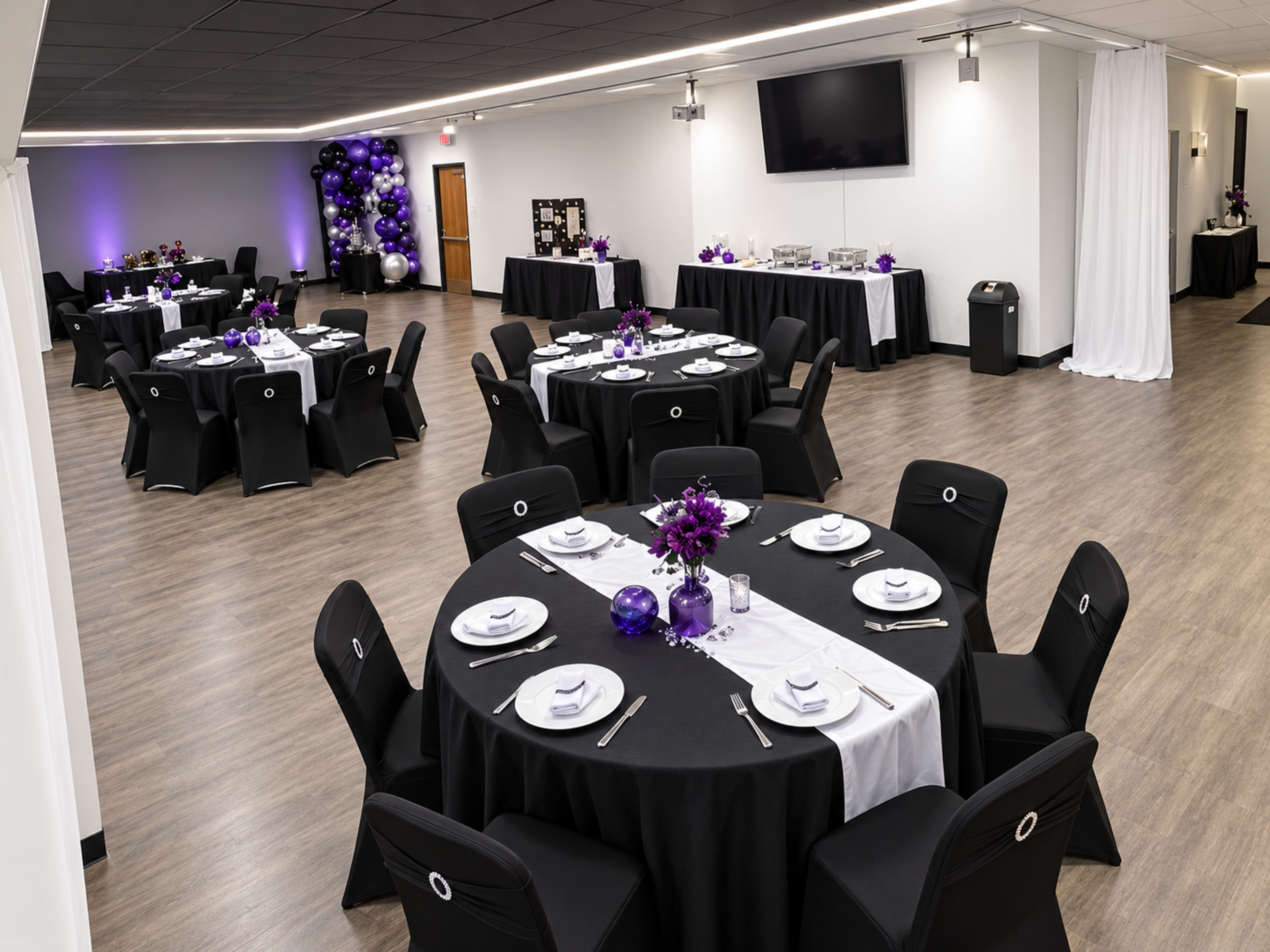 A spacious event room features round tables covered with black tablecloths and set with white plates and purple centerpieces, while purple balloons adorn one corner.