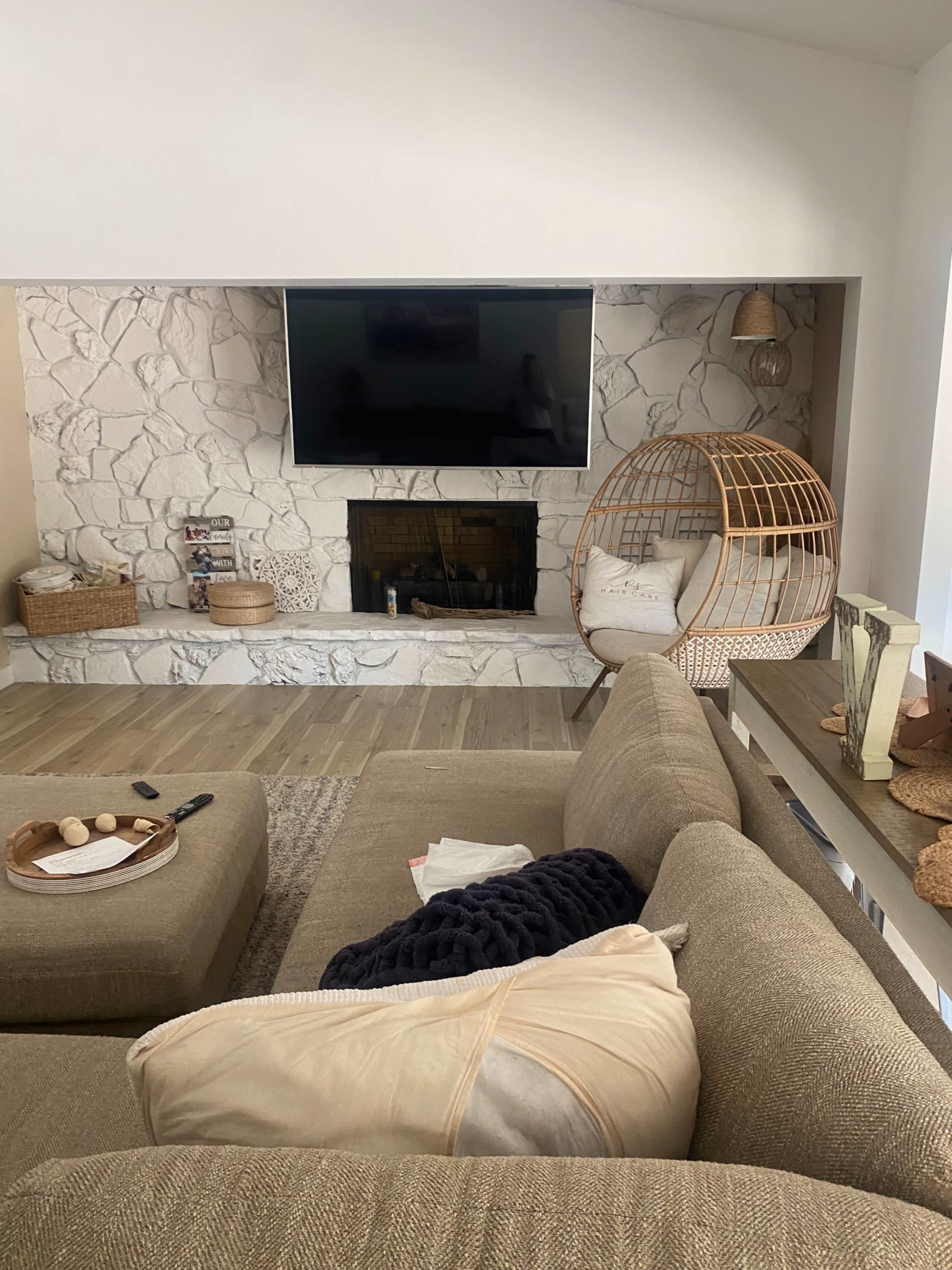 The image shows a modern living room featuring a large television mounted above a stone fireplace, a woven hanging chair, and a sectional sofa with neutral-colored cushions.