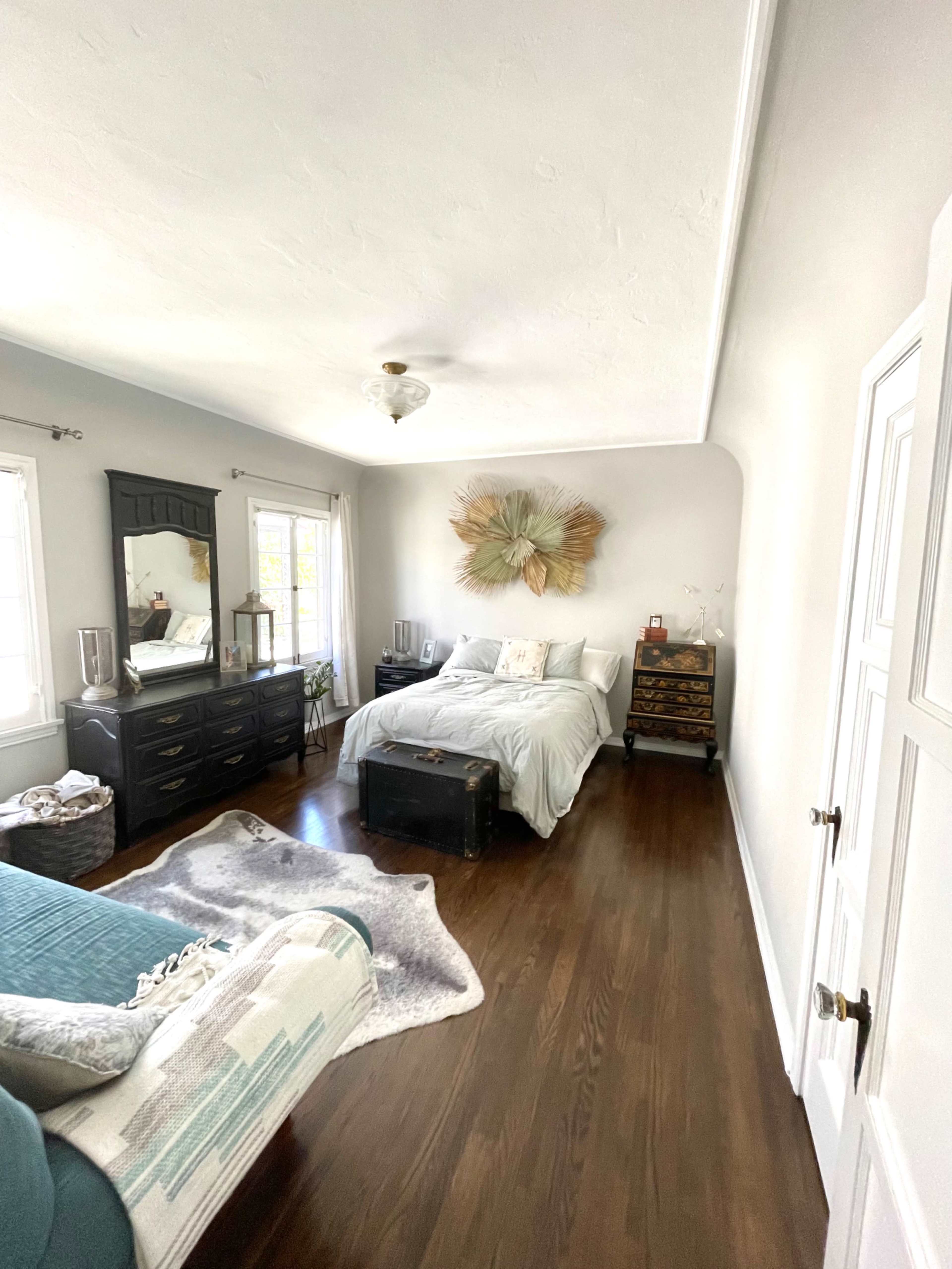 The image shows a bright bedroom featuring a bed with light bedding, a dark wooden dresser, and a decorative piece made of leaves on the wall.