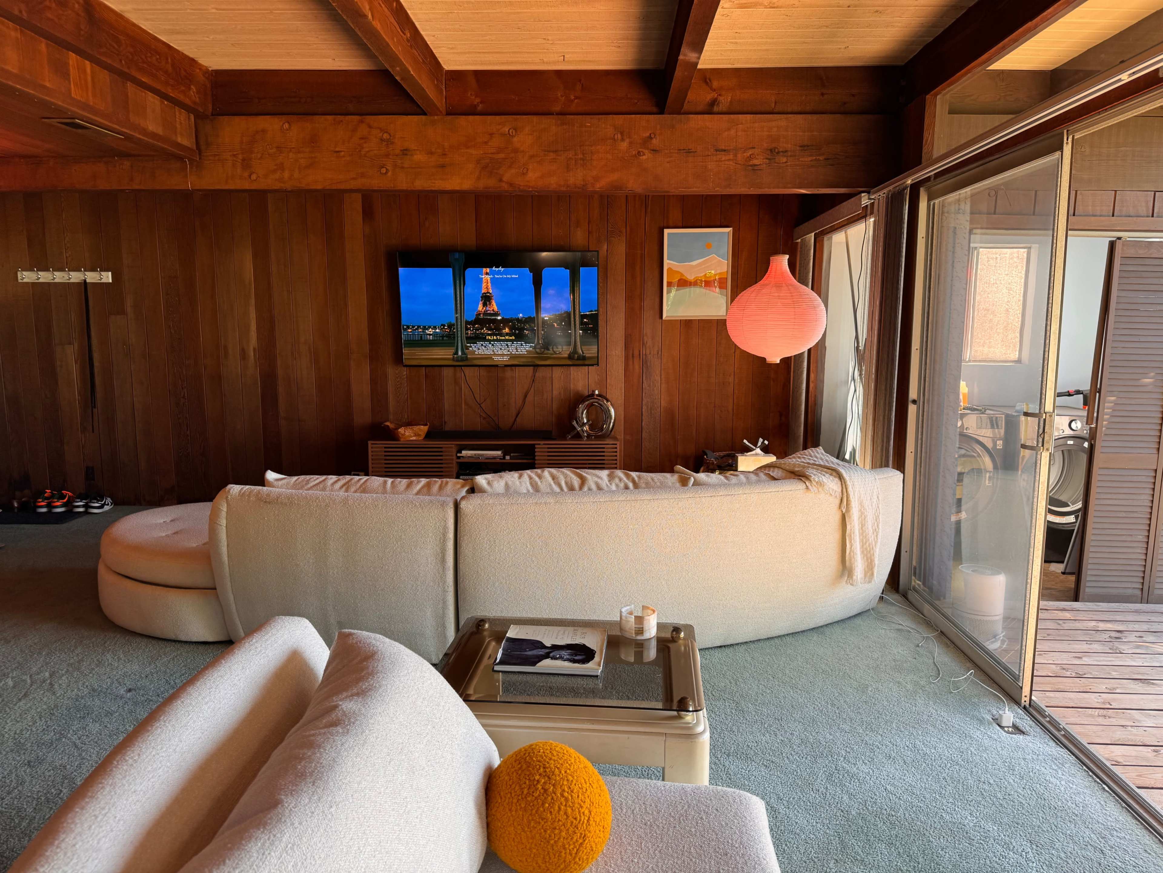 A modern living room with a curved white sofa, a television displaying an Eiffel Tower image, wooden walls, and large glass doors leading to a deck.